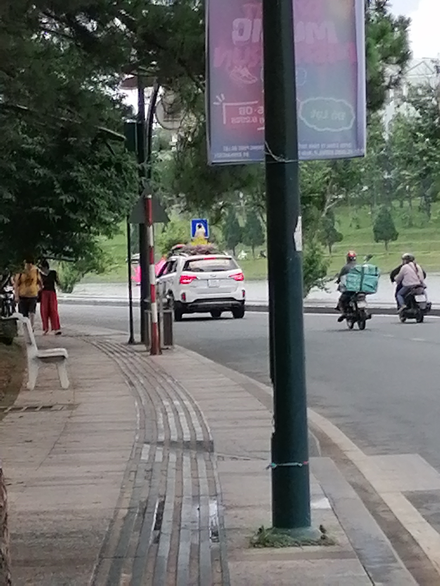 The scene is set in Da Lat, Vietnam, with a sidewalk on the left and a road on the right. The sidewalk has tactile paving for the visually impaired. There's a white park bench and two people walking on the sidewalk. A white SUV is on the road with something on its roof rack, possibly plants. Two people on scooters are also on the road. In the foreground is a pole with a sign. The environment appears to be a green, urban area.