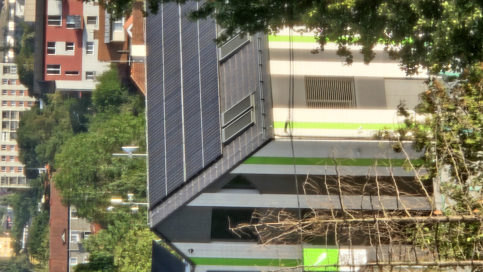 The image shows a modern building with solar panels on its roof, situated in a residential area of Prešov, Slovakia. The building features a distinctive design with vertical green and white stripes on its facade. In the background, other residential buildings and lush green trees are visible, suggesting a suburban or semi-urban setting. The overall impression is one of sustainable urban development in a well-treed environment.