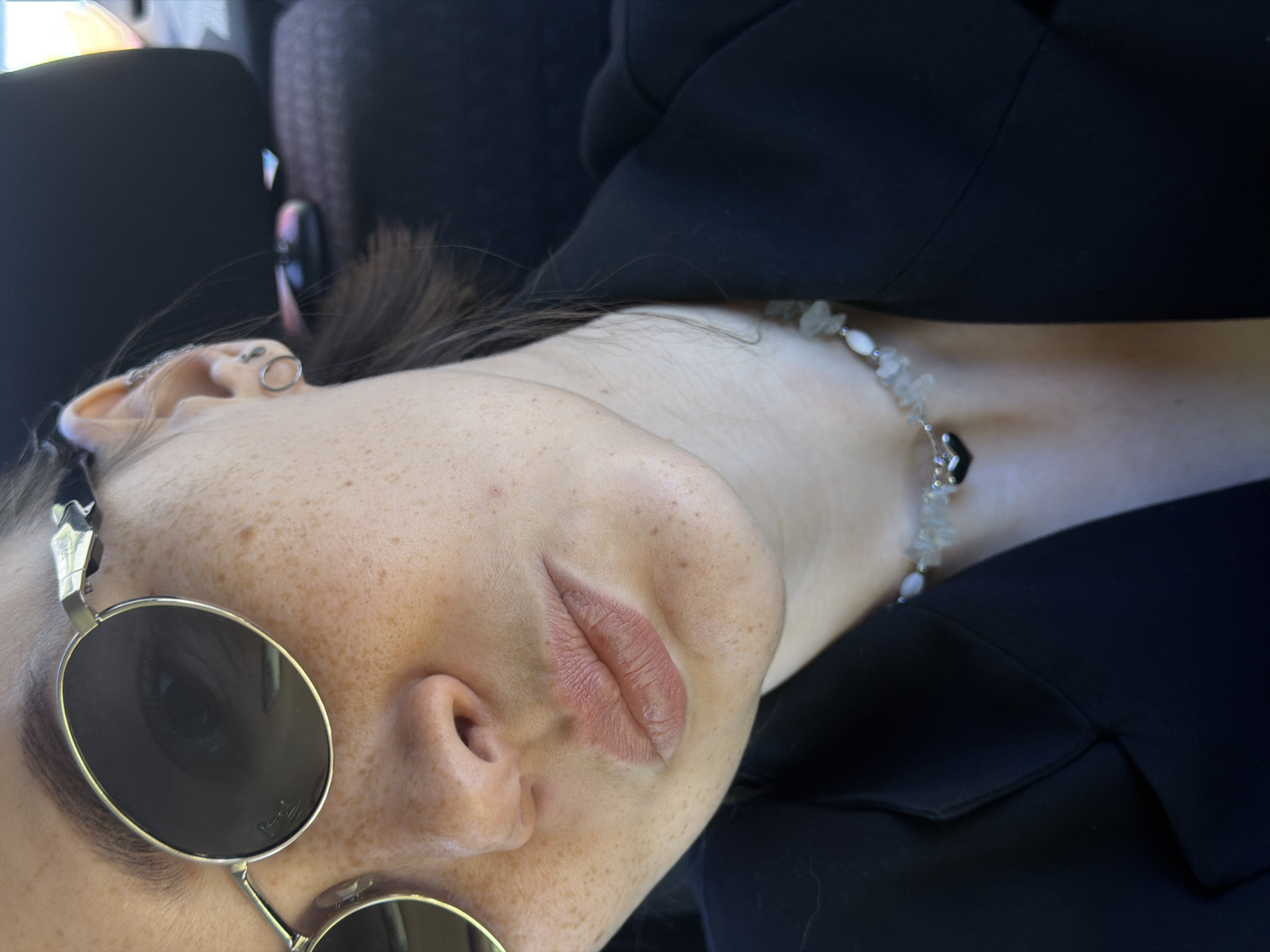 A close-up, angled shot depicts a person with freckled skin, wearing round sunglasses with a gold frame. Only one eye is visible behind the tinted lens. The person has an ear piercing and a necklace with light-colored stones and a black pendant around their neck. They are wearing a black blazer or coat. The background includes a dark headrest, possibly inside a car. The photo does not provide enough information to confirm the location in the city of Chaikovsky, Russia.