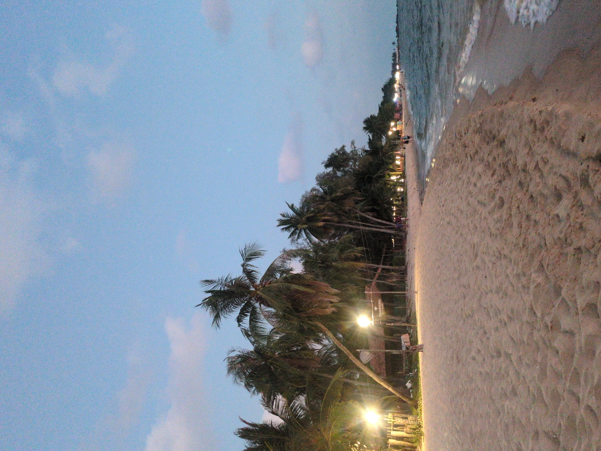 The image depicts a beach scene at night in Phú Quốc, Vietnam. The sky is a dusky blue with scattered clouds. A line of palm trees and vegetation separates the beach from a walkway lined with lights. The sand is light-colored and extends to the water's edge, where gentle waves are visible. A few people are visible in the distance, walking along the walkway. There is a sign for "THANHKRIEU".