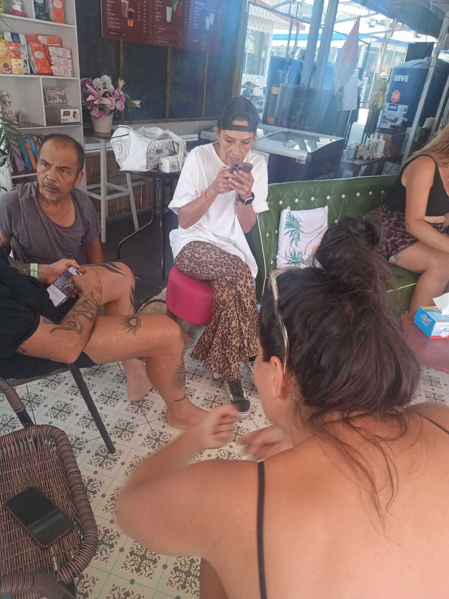 The image shows a group of people sitting in a casual setting, possibly a cafe or restaurant in ตำบลเกาะพะงัน, Thailand. An older Asian man is seated to the left, looking off-camera. Another person with tattoos is using a phone, and a woman wearing a white t-shirt and leopard-print pants is also using a phone. Two other women are partially visible. The scene includes furniture like chairs, a sofa, and a small table. The environment suggests an indoor location with access to an outdoor area.