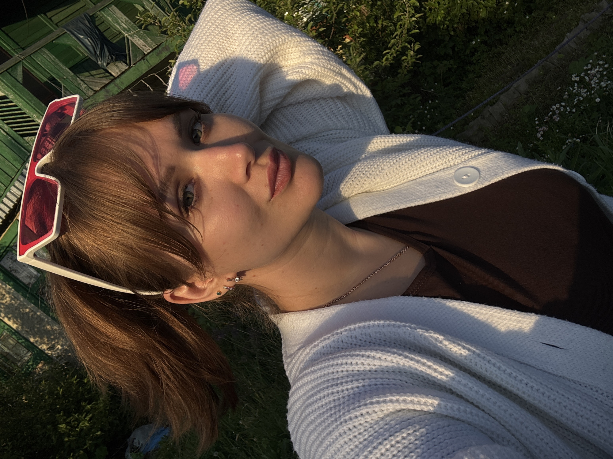 A young woman is depicted in a close-up, eye-level shot, appearing relaxed and lying down outdoors in what seems to be a grassy area or garden. She has medium brown hair, pulled back into a ponytail with some bangs framing her face. She is wearing a light-colored (possibly white or off-white) knitted cardigan over a dark brown top. Red and white-framed sunglasses are pushed up onto her head. Her face is turned slightly towards the viewer, with a soft, calm expression. Notable facial features include her light-colored eyes and full lips. She wears multiple earrings in her left ear, some with small gems, and a thin chain necklace.
The lighting suggests late afternoon or early evening, often referred to as "golden hour," with warm, directional light creating distinct shadows on her face, hair, and clothing, highlighting textures. The background shows lush green foliage, possibly bushes and grass with small white flowers, indicating a natural outdoor setting. To the left, there's a structure with green wooden slats, possibly a fence or part of a building, and some dark fabric draped over it.
The overall scene conveys a peaceful and relaxed moment. There is no visible text in the image. The location context provided is Chaikovsky, Russia.