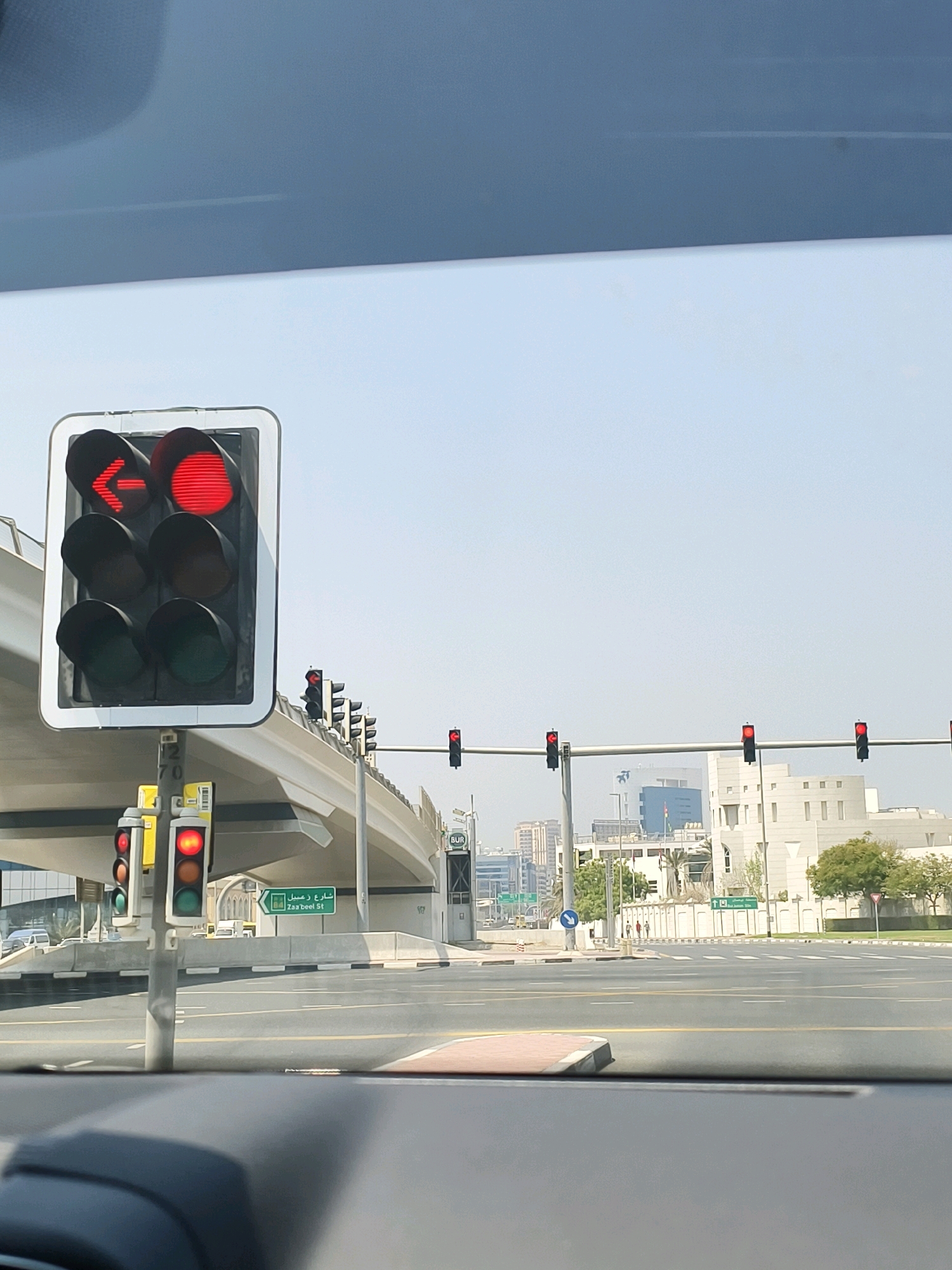 This image captures a view from a vehicle on a street in Dubai, United Arab Emirates, under a clear, bright sky. In the foreground, a traffic light pole with multiple signals is visible. The primary traffic signal displays a red left-turn arrow and a solid red circle. Below this, another traffic signal shows a red light on top and an amber light in the middle. Further down the road, a series of traffic lights hang across the intersection, all illuminated with red lights. A large overpass or bridge arches over the intersection. A green street sign with Arabic script and "Za'abeel St" written in English is mounted on a pole to the right of the primary traffic light. In the background, modern buildings, including a tall skyscraper with a blue facade, form part of the Dubai skyline. Trees and landscaping are present along the sides of the road. The road itself is paved with asphalt and marked with white and yellow lines, indicating multiple lanes. Several vehicles are either parked or in motion in the distance. The overall scene suggests a typical urban traffic environment in Dubai.