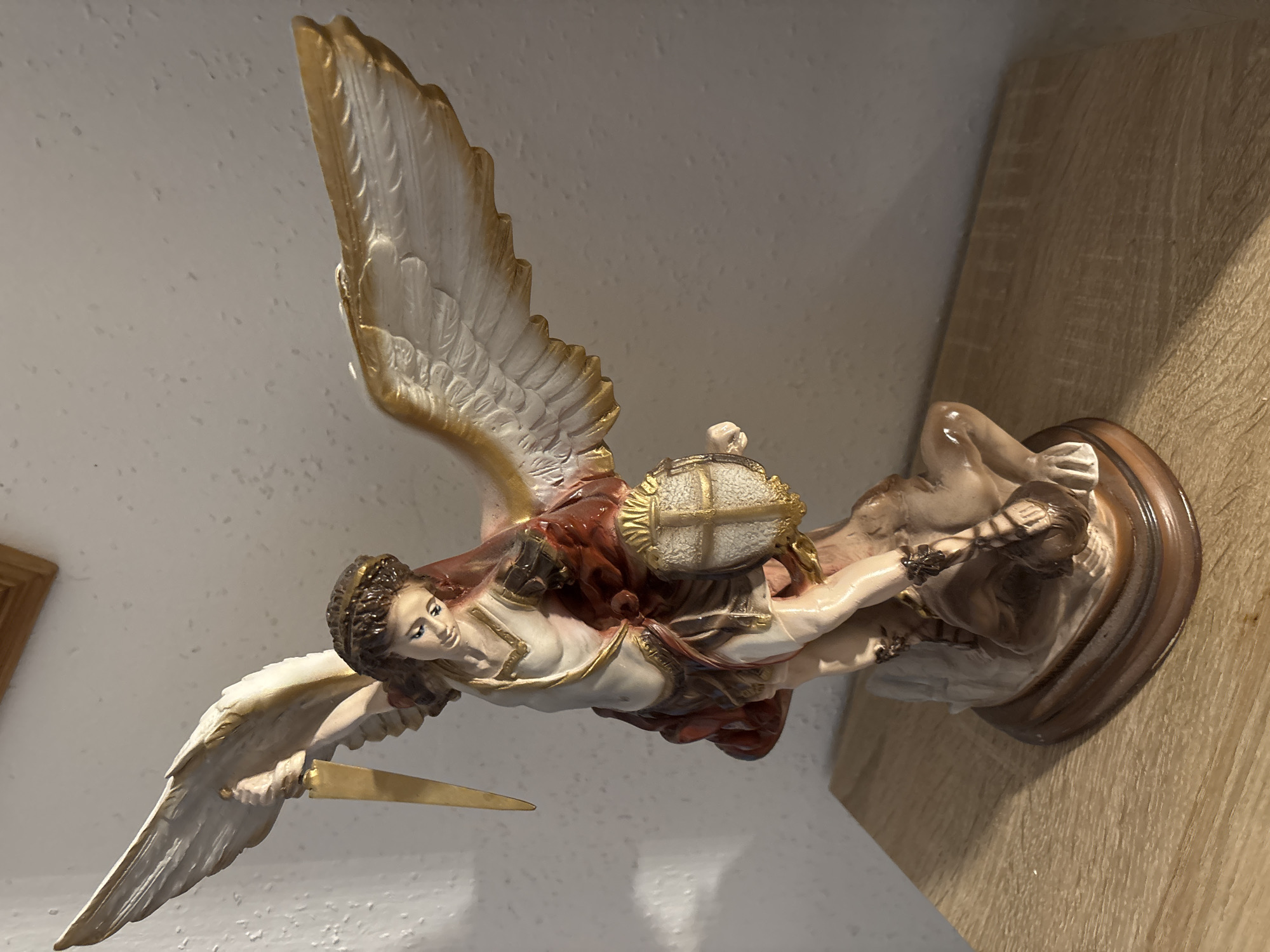 A statue of Saint Michael is displayed on a wooden surface. The statue depicts Saint Michael with wings, holding a sword in his right hand and a shield with a cross emblem in his left. He is shown triumphing over a beast at his feet. The background is a textured wall. This scene is not located in the city of Celle, Germany.