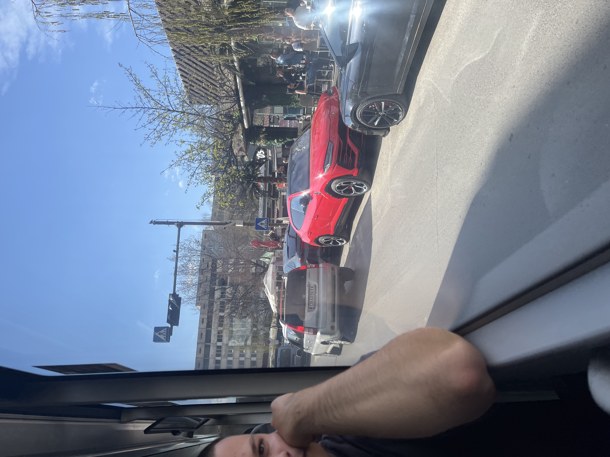 The image captures a street scene in Sofia, Bulgaria, viewed from inside a vehicle. Visible in the foreground is part of a person's arm and face within the car. Through the car's window, the street presents a bright daylit scene with a blend of urban elements. Several cars are parked along the side of the road, including a red sports car prominently positioned in the middle of the frame, as well as a silver van. There are people sitting at tables outside a café or restaurant. Buildings line the street, adding to the urban setting. A pedestrian crossing sign is visible.