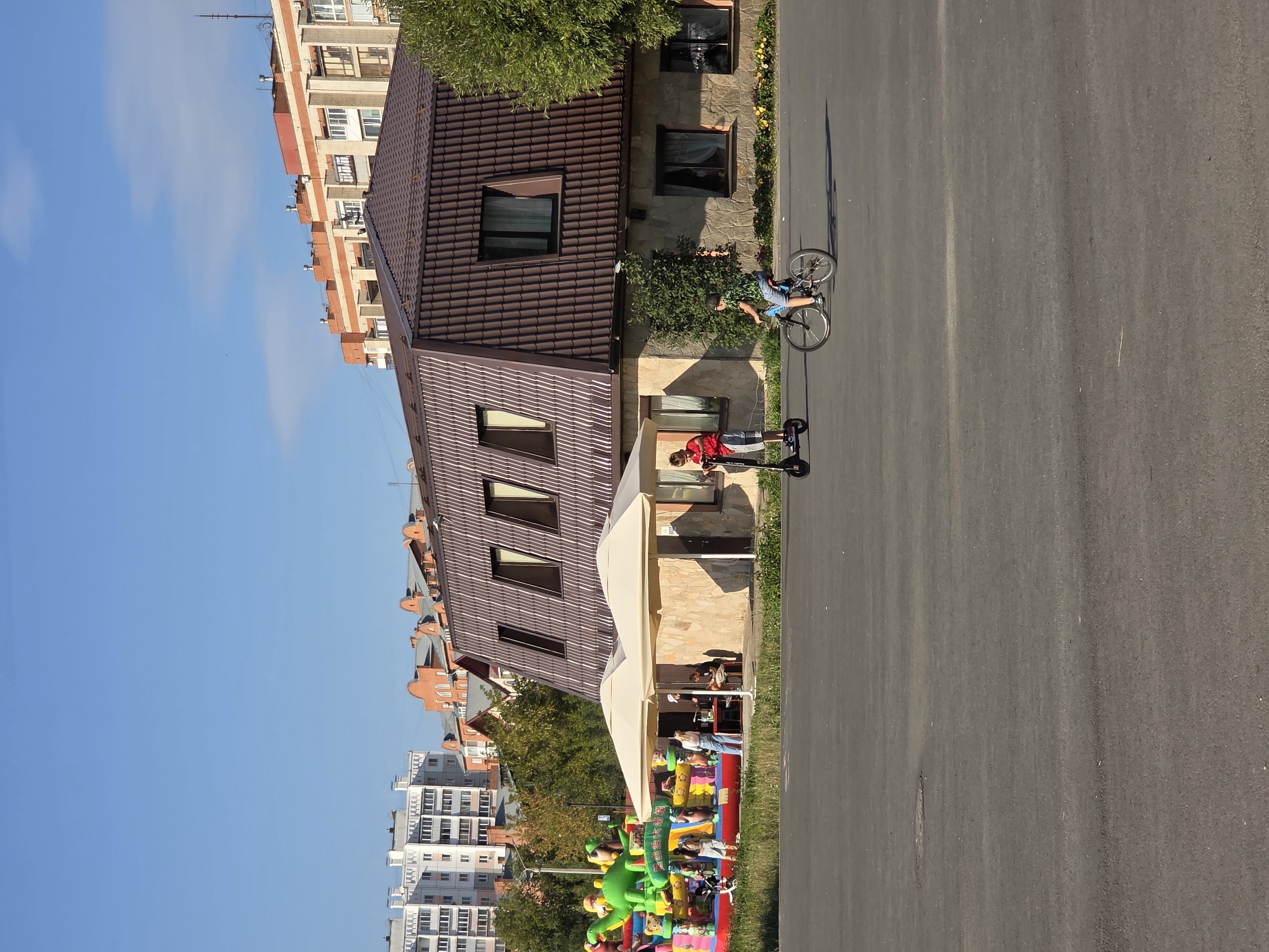 The image depicts an outdoor scene on a sunny day in Miass, Russia. In the foreground, a paved area is visible, with a man on a bicycle to the right and a boy on an electric scooter to the left. Behind them is a building with a brown, multi-gabled roof and stone-colored walls. In front of this building, there are two large, cream-colored umbrellas providing shade over an outdoor seating area where several people are gathered. To the far left of the frame is a large, inflatable playground structure with various cartoon characters. In the background, multiple residential buildings of varying architectural styles are visible, along with trees and a clear blue sky. The scene appears to be a public space, possibly a park or a recreational area adjacent to a cafe or restaurant.