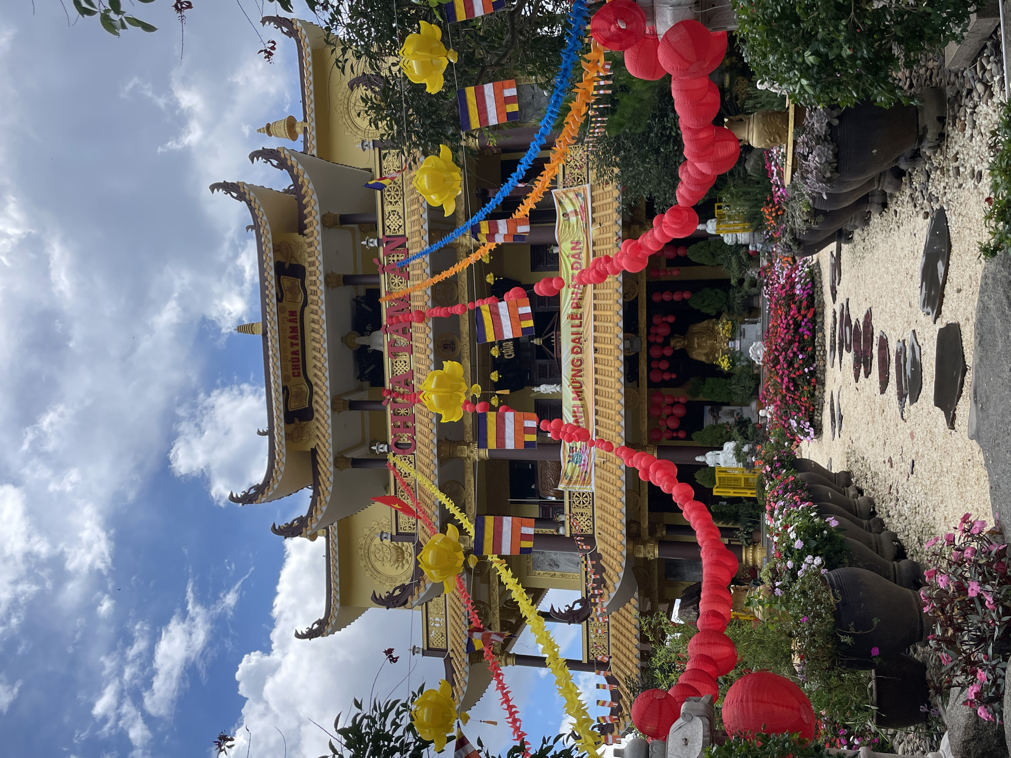 This media file depicts a Buddhist temple in Da Lat, Vietnam, likely during a festival or celebration. The primary subject is the ornate, yellow temple building with tiered roofs and intricate architectural details. The foreground is adorned with strings of red paper lanterns and colorful paper chains in yellow and blue. Small, multi-colored flags, possibly representing Buddhist denominations or national flags, are also visible.
The scene is bathed in bright daylight under a partly cloudy sky, suggesting a pleasant day. Lush greenery and vibrant flowers, including pink and purple blossoms, surround the temple grounds, adding to the festive atmosphere. Large, dark ceramic pots are strategically placed, some containing plants. A pathway paved with light-colored gravel and dark stone slabs leads towards the temple entrance.
Although no people are clearly visible in the foreground or mid-ground, the festive decorations imply a gathering or event is taking place or has recently concluded. The overall emotion conveyed is one of celebration and spiritual significance.
Visible text on a banner above the temple entrance reads "MỪNG ĐẠI LỄ PHẬT ĐẢN" which translates to "Happy Buddha's Birthday," confirming the event. The presence of Buddhist iconography, such as a large golden Buddha statue visible in the background and decorative symbols on the temple, further anchors the location as a religious site. The architecture and decorations are characteristic of Vietnamese temples, particularly those found in tourist destinations like Da Lat.