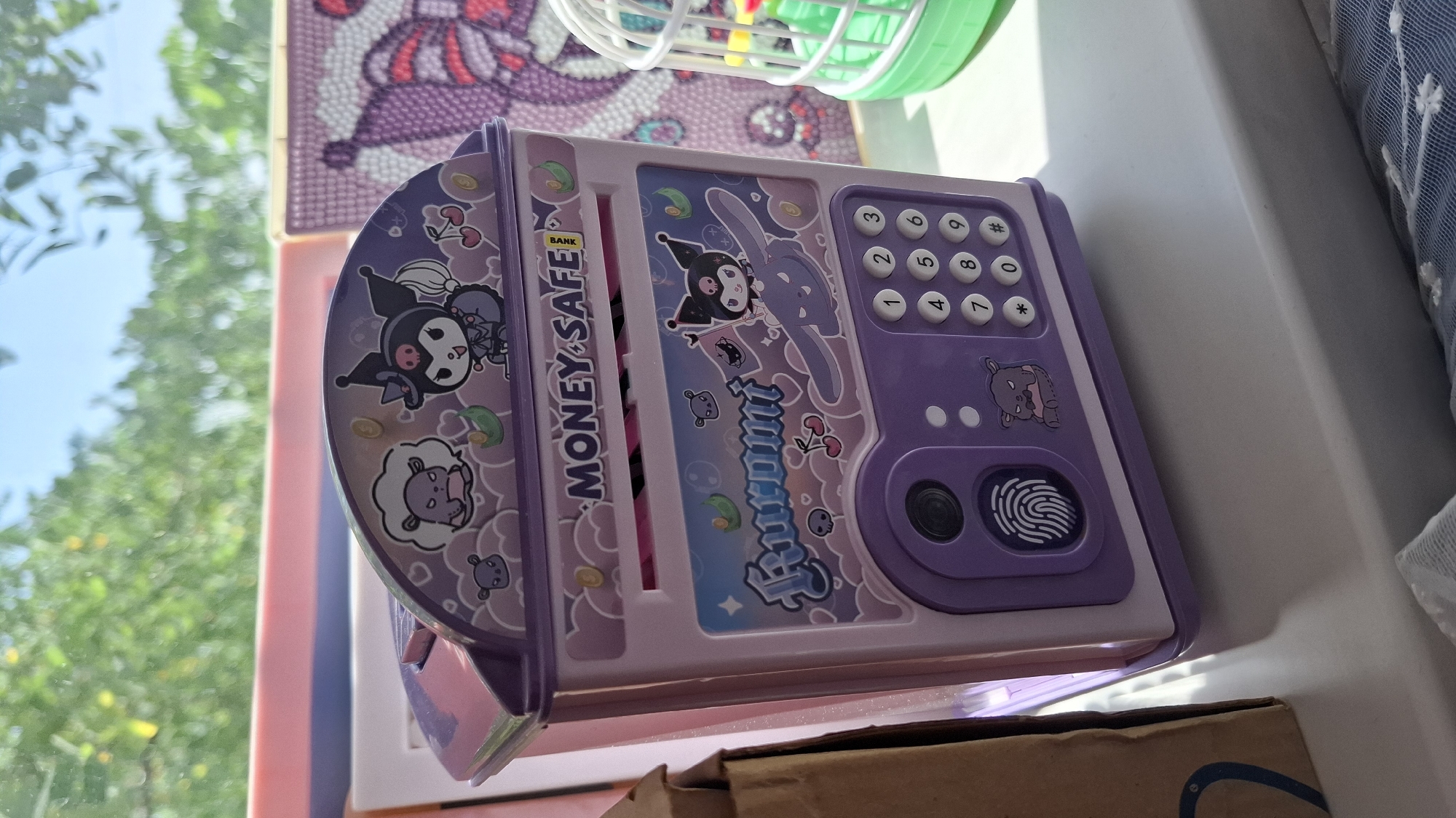 A close-up, eye-level, outdoor shot captures a cartoon-themed purple and pink money safe sitting on a windowsill, with a background of blurred green foliage and blue sky, suggesting it is daytime. The safe features colorful decals of cartoon characters, including one resembling "Kuromi" from Sanrio, a fingerprint scanner, and a numeric keypad. To the left, a brown cardboard box is partially visible. To the right, a white birdcage with a green base contains colorful toys. The scene is set on what appears to be a windowsill in Shymkent, Kazakhstan. The image is sharp and well-lit, with natural light illuminating the objects.