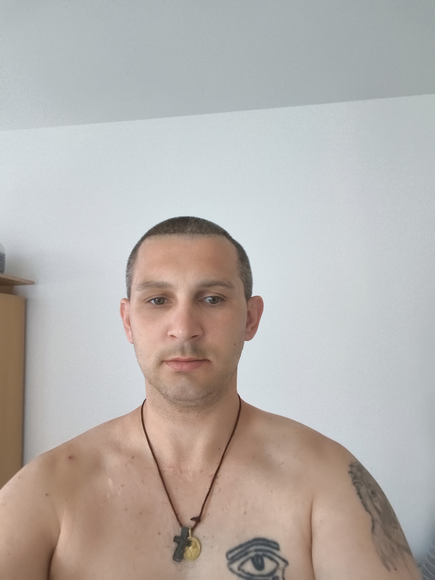 A man with a shaved head and light brown eyes looks directly at the camera. He is shirtless, wearing a necklace with a cross and a round pendant. He has a tattoo of an eye on his chest and another tattoo on his right shoulder. His skin shows some discoloration and scarring on his neck and upper chest. In the background, a light-colored wall and a wooden cabinet are visible. The image appears to be taken indoors, possibly in a residential setting in Wolgast, Germany.