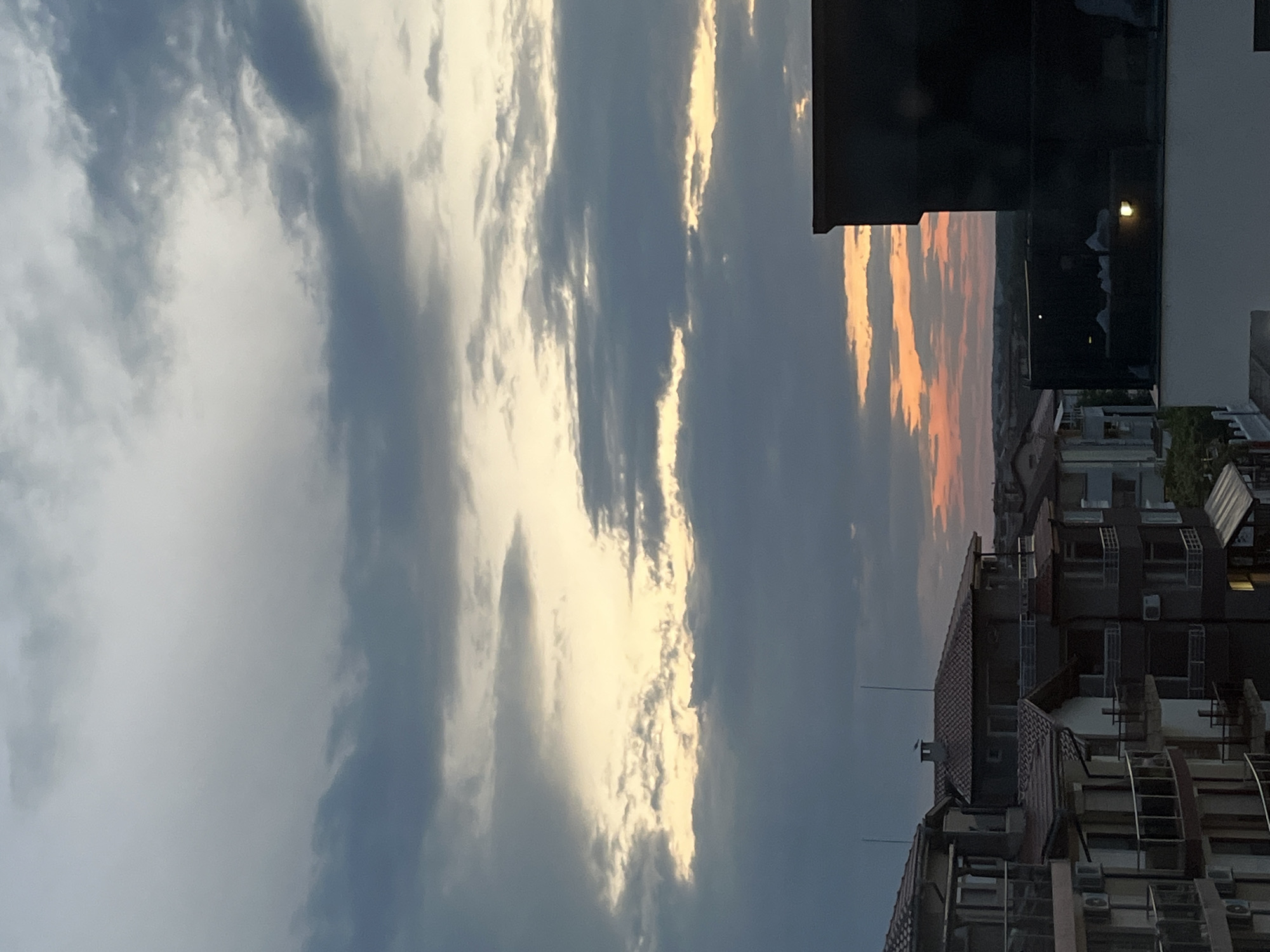 An aerial view captures the twilight sky over Lozenets, Bulgaria, casting a soft glow on a residential area. The sky is dominated by a dramatic arrangement of clouds, with darker, heavier formations in the upper portion gradually giving way to lighter, wispy clouds illuminated by the setting sun. Patches of warm orange and yellow peek through the cloud cover, indicating the horizon lies beyond the frame.
In the foreground, apartment buildings are visible, their tiled roofs and balconies arranged in a layered fashion. The architecture suggests a modern residential development. The buildings are a mix of light and dark facades, with visible windows and air conditioning units. The lower section of the image is partially obscured by the dark silhouette of a rooftop structure or a balcony railing, adding a sense of depth to the scene. A faint light source is visible on this structure. The overall impression is one of a quiet evening settling over the town.