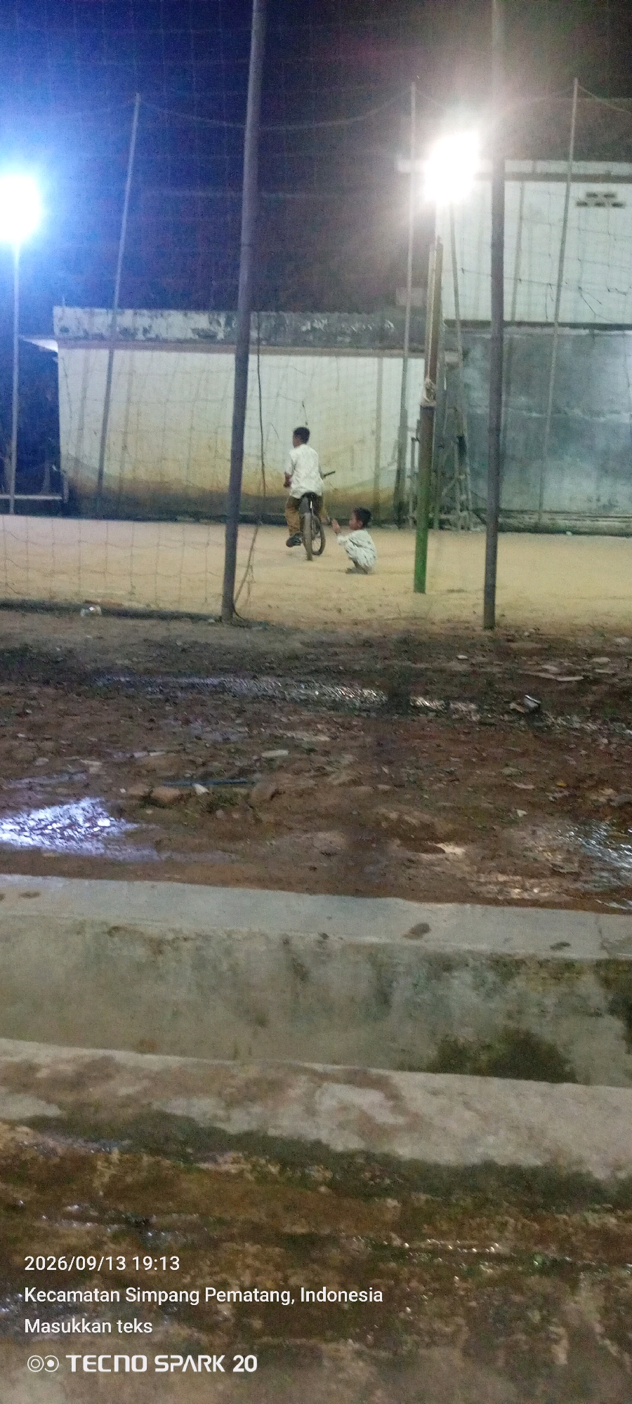 The image depicts an outdoor scene at night, illuminated by artificial lights, likely at a sports court or a similar open area. Two young individuals are present. One child is seated on the ground, facing towards the right. Another child is riding a bicycle, moving from left to right across the frame. In the background, a light pole stands vertically, and beyond that, a weathered wall with a net structure is visible. The ground appears to be dirt or sand, with some patches of water reflecting the light. The lower portion of the image shows a concrete structure. Text overlays indicate the date and time as "2026/09/13 19:13," and the location as "Kecamatan Simpang Pematang, Indonesia." A logo for "TECNO SPARK 20" is also present.
