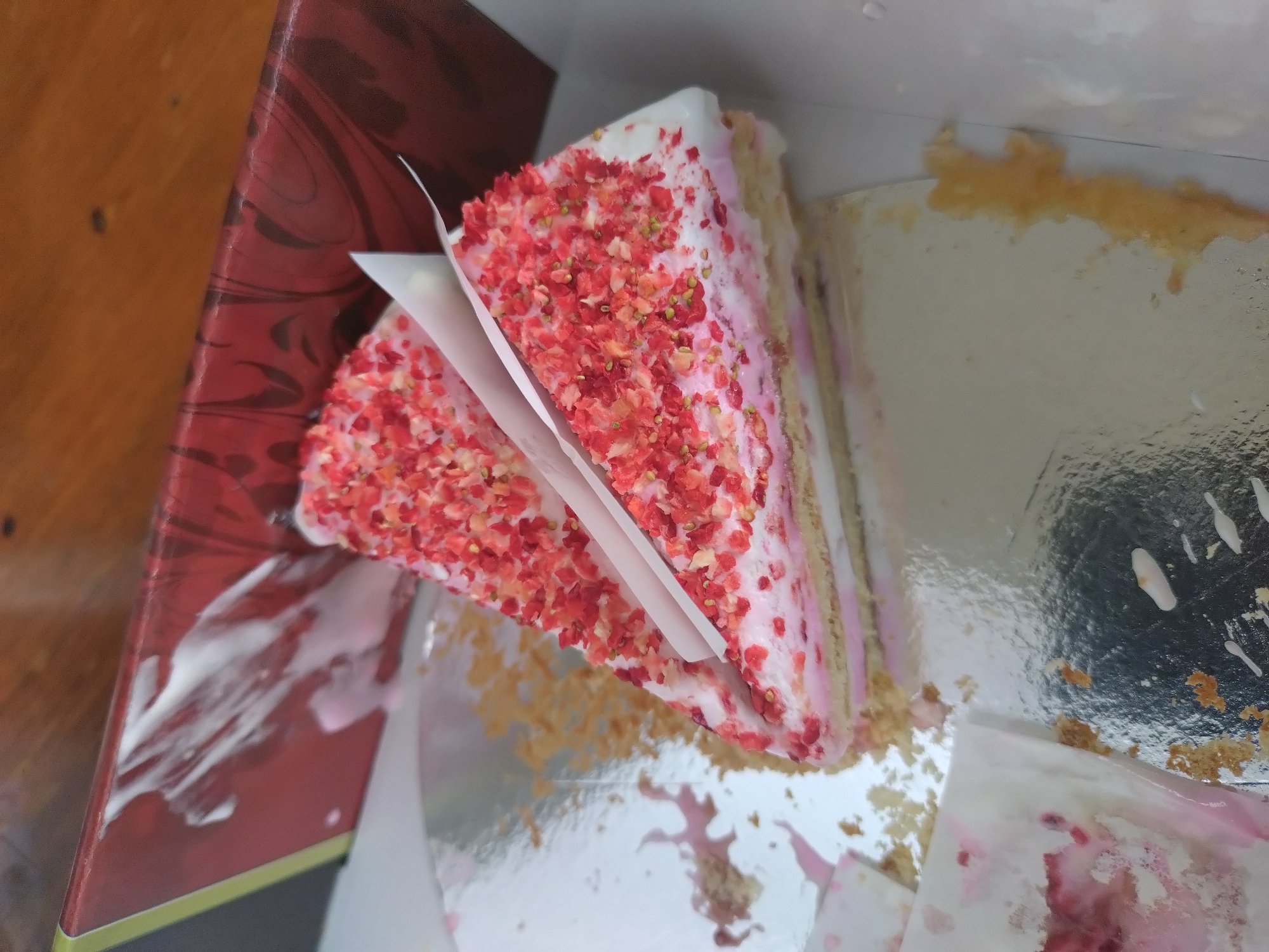 A triangular slice of layered cake, topped with white frosting and pink strawberry crumble, rests on a silver platter. The cake appears to be partially eaten, with crumbs scattered on the platter and a small piece of white paper inserted into the slice. A red, decorative box is visible in the background, out of focus. The scene suggests a dessert served at a dining table. The location is Yagoda, Bulgaria.