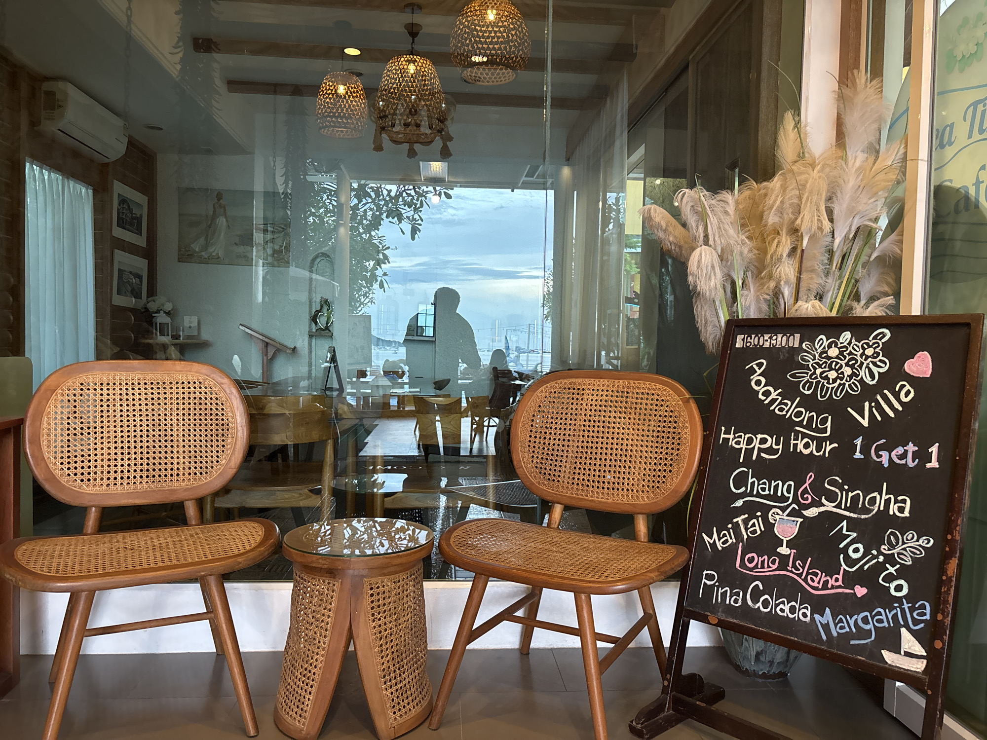 The image depicts an outdoor seating area of a cafe or restaurant in Chalong, Thailand, with large windows reflecting the interior and offering a view of the outdoors. In the foreground, two intricately woven rattan chairs with wooden frames face a small, similarly constructed side table with a glass top. To the right, a chalkboard sign displays a menu with handwritten drink names and prices, including "Happy Hour 1 Get 1 Chang & Singha," "Mai Tai," "Long Island," "Mojito," "Pina Colada," and "Margarita." The sign also features decorative drawings of flowers and a small sailboat. Behind the chairs and sign, the interior of the establishment is visible through the glass, showing more seating, decorative lights hanging from the ceiling, and framed pictures on the wall. The outdoor view through the windows reveals a cloudy sky and a silhouette of a person standing near a window, with a body of water and boats visible in the distance. The overall atmosphere suggests a relaxed, tropical setting.