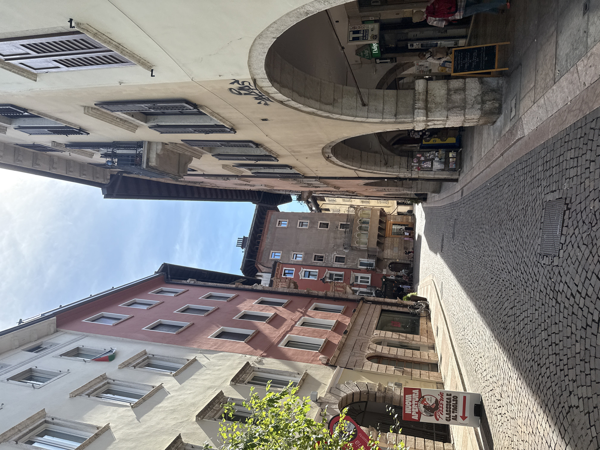 This is an exterior, eye-level, daytime, vertical shot of a narrow, cobblestone street in the historic center of Trento, Italy. The street is lined with old, multi-story buildings constructed of stone and plaster. The buildings on the left are painted in pastel colors and have many windows with shutters. The building on the right is a lighter color and features arched walkways on the ground floor, with shops or cafes visible behind the arches. There are people walking and standing in the street and under the arches. A red and white sign for "NUOVA APERTURA PIZZERIA" is visible on the left, indicating a new pizza place. The sky is blue with some clouds, and sunlight casts shadows across the cobblestones.