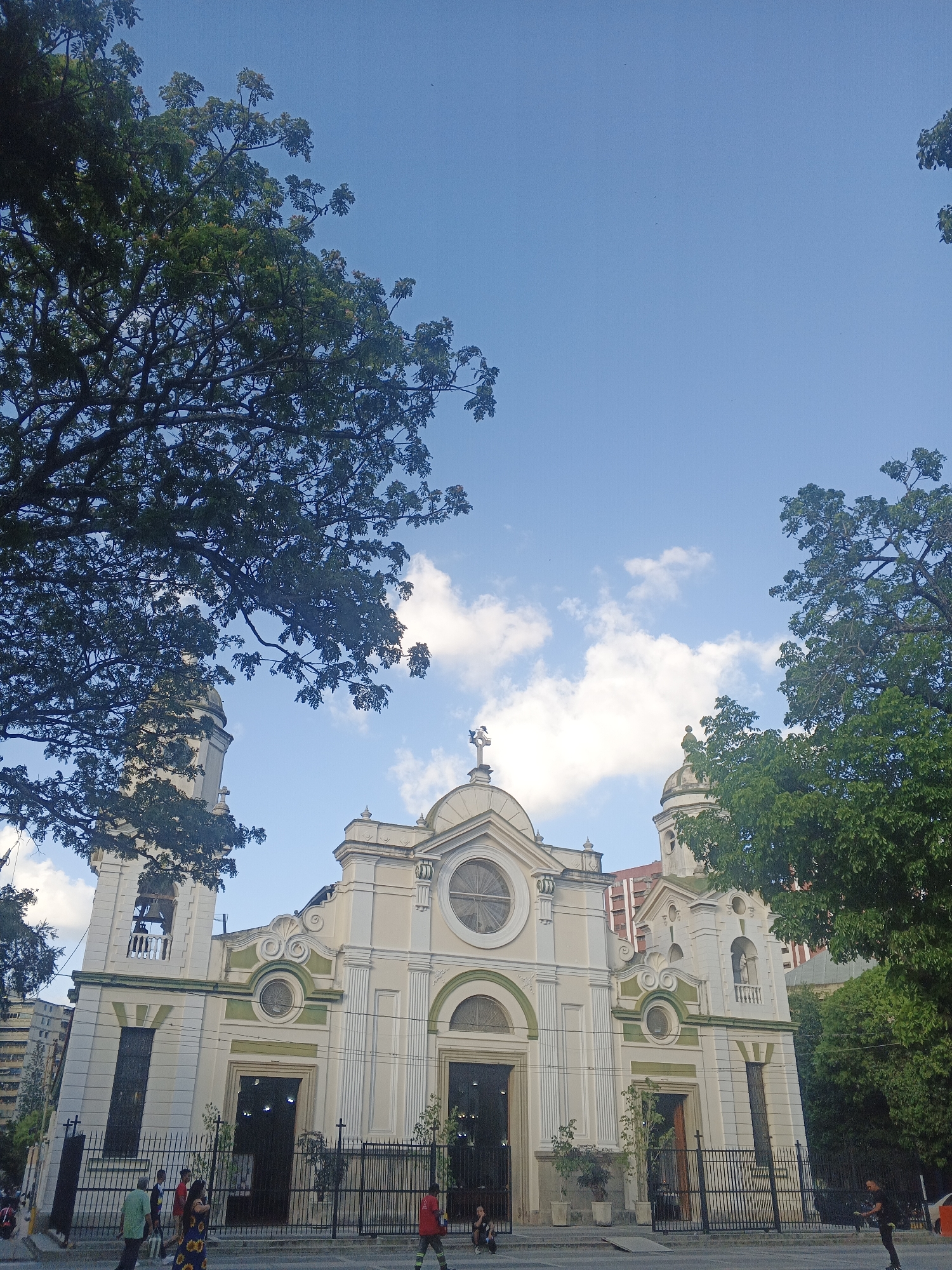 The image captures a low-angle view of a church facade in Caracas, Venezuela, on a bright day. The church is painted in shades of off-white, cream, and light green, featuring two bell towers on either side. A prominent cross sits atop the central architectural feature above the main entrance. The sky is blue with scattered white clouds. Trees with dense foliage frame the church on either side, partly obscuring the view. People are visible near the church entrance, standing and walking. A black wrought-iron fence runs along the front of the church. A tall building can be seen behind the church.