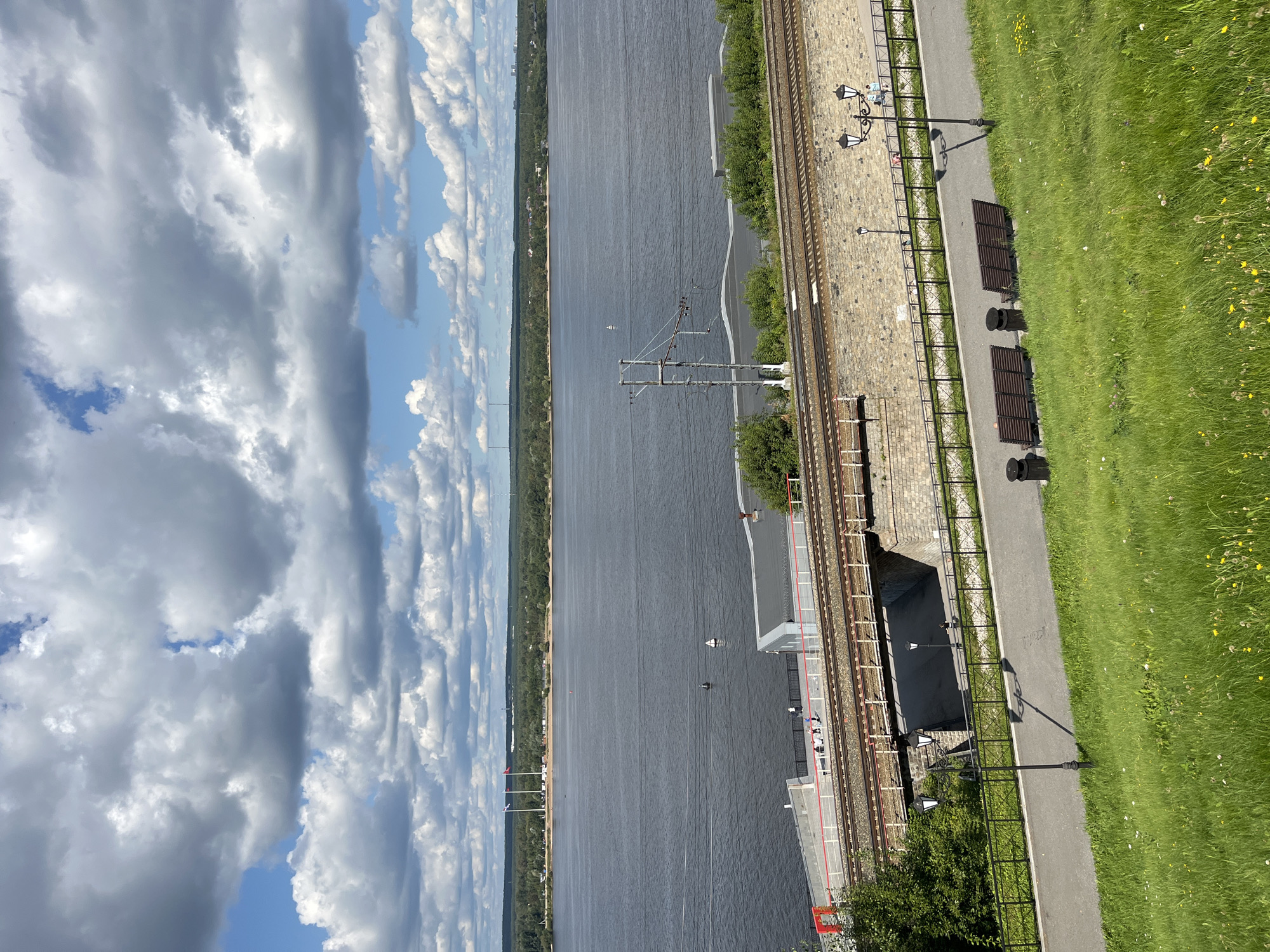 The image captures a scenic view of the Kama River in Perm, Russia, from an elevated perspective. In the foreground, a lush green grassy slope dotted with small yellow flowers slopes down towards a paved walkway. Along the walkway, there are several dark wooden benches and trash receptacles. Two ornate lampposts cast shadows on the path.
Behind the walkway, a stone embankment supports a set of railway tracks. A bridge carrying the tracks spans over a lower area. Beyond the embankment and tracks, a body of water, the Kama River, stretches out. On the far bank of the river, a treeline forms a dense green horizon, with some distant buildings and what appear to be industrial structures or towers visible. The sky above is filled with dramatic, large cumulus clouds against a backdrop of blue sky. A few buoys or markers are visible in the river. People can be seen walking along the walkway and on the embankment near the railway tracks. The overall atmosphere is serene and picturesque.