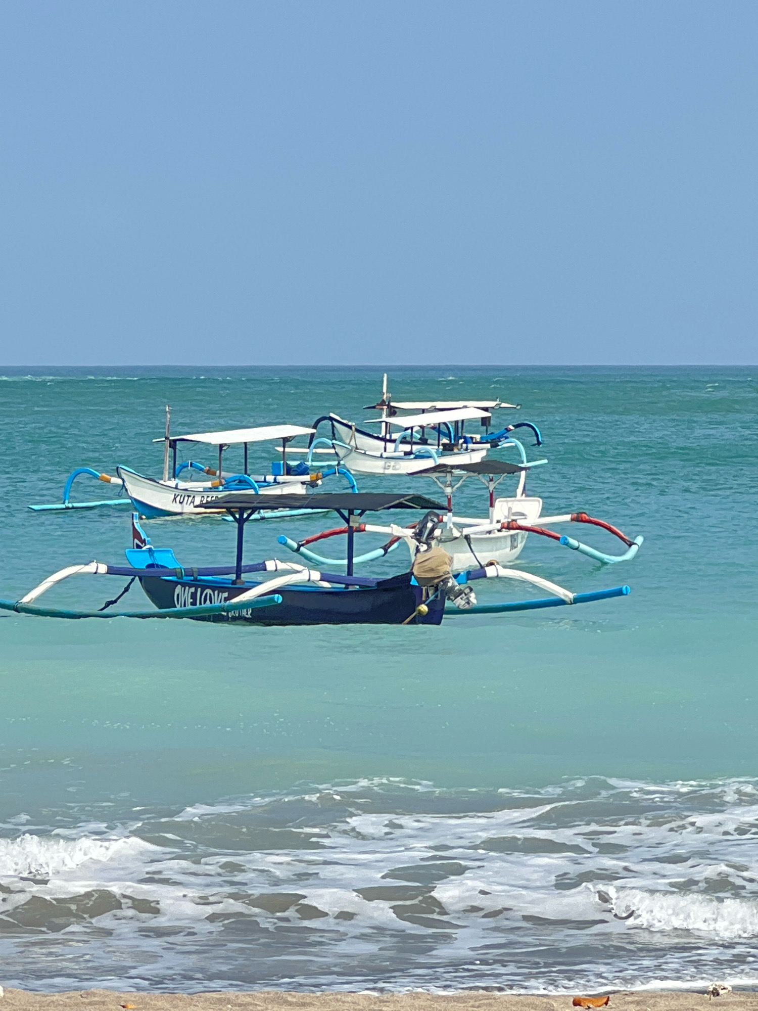 Two traditional outrigger fishing boats are anchored in the turquoise waters off the coast of Kuta, Indonesia. The boat in the foreground is dark blue with the words "ONE LOVE" written on its side in white lettering, and the word "KUTA" is visible in white on the white portion of the boat behind it. Both boats have white canopies supported by poles and colorful outriggers extending from their sides. The sea is calm with gentle waves approaching the sandy shore in the foreground. The sky is a clear, pale blue.