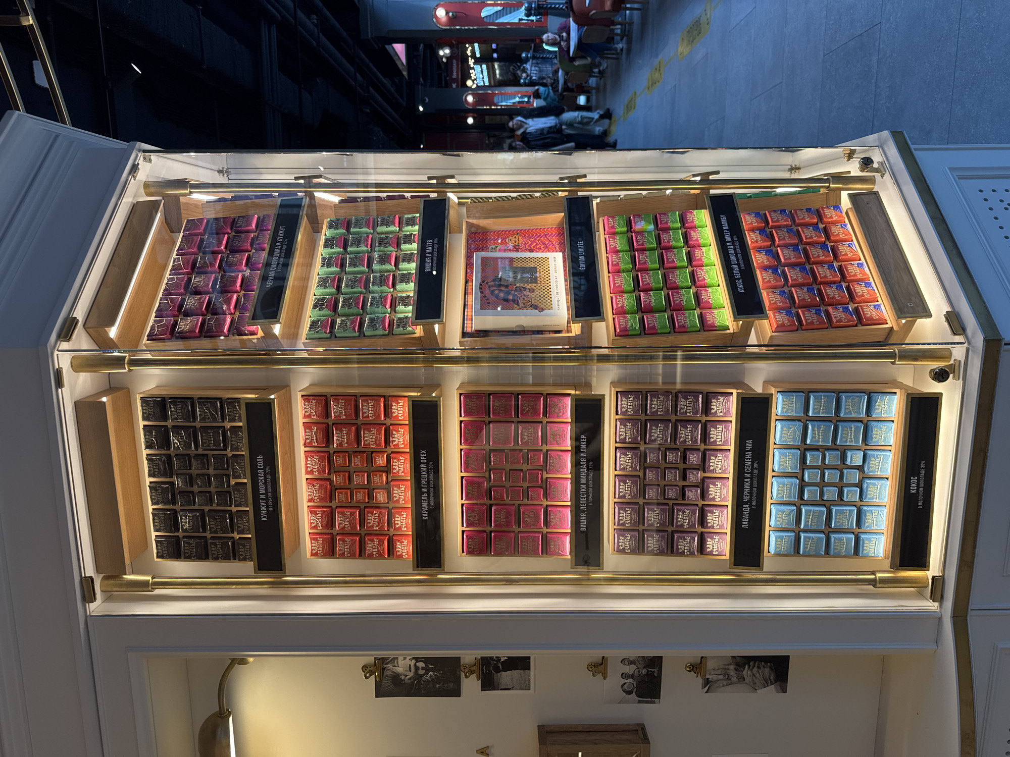 The image, rotated clockwise by 90 degrees, presents a brightly lit display case showcasing an assortment of individually wrapped gourmet chocolates. The setting is indoors, likely a confectionery store or cafe in Saint Petersburg, Russia, given the Cyrillic text.
The elegant display case features a white frame, gold-colored metal accents, and wooden dividers, organizing the chocolates into multiple tiered compartments. Each small, square chocolate is uniformly wrapped in foil, bearing the Russian word "СЧАСТЬЕ" (Schastye), meaning "Happiness."
In the top row, from left to right:
- Dark purple/magenta chocolates are labeled "ЧЕРНАЯ СМОРОДИНА И КУНЖУТ В ГОРЬКОМ ШОКОЛАДЕ 72%" (Blackcurrant and Sesame in Dark Chocolate 72%).
- Green chocolates are identified as "ВИШНЯ И МАТТЯ • МОЛОЧНОМ ШОКОЛАДЕ 30%" (Cherry and Matcha in Milk Chocolate 30%).
- A central section holds a special "ÉDITION LIMITÉE" (Limited Edition) box with vibrant artwork.
- Another section of green chocolates appears, similar to the previous green ones, without a unique label clearly visible.
- Red/orange chocolates are labeled "КОКОС, БЕЛЫЙ ШОКОЛАД И ЛИКЕР МАЛИБУ" (Coconut, White Chocolate and Malibu Liqueur).
The bottom row features:
- Dark brown/black chocolates, labeled "КУНЖУТ И МОРСКАЯ СОЛЬ В ГОРЬКОМ ШОКОЛАДЕ 72%" (Sesame and Sea Salt in Dark Chocolate 72%).
- Red/orange chocolates, identified as "КАРАМЕЛЬ И ГРЕЦКИЙ ОРЕХ В МОЛОЧНОМ ШОКОЛАДЕ 30%" (Caramel and Walnut in Milk Chocolate 30%).
- Pink chocolates, labeled "ВИШНЯ, ЛЕПЕСТКИ МИНДАЛЯ И ЛИКЕР В ГОРЬКОМ ШОКОЛАДЕ 72%" (Cherry, Almond Petals and Liqueur in Dark Chocolate 72%).
- Purple chocolates, identified as "ЛАВАНДА, ЧЕРНИКА И СЕМЕНА ЧИА В МОЛОЧНОМ ШОКОЛАДЕ 30%" (Lavender, Blueberry and Chia Seeds in Milk Chocolate 30%).
- Light blue chocolates are simply labeled "КОКОС В МОЛОЧНОМ ШОКОЛАДЕ 30%" (Coconut in Milk Chocolate 30%).
The background, out of focus, shows a bustling public area, possibly a mall concourse or street, with several people walking and sitting at tables. The overall atmosphere is well-lit, suggesting daytime. Below the chocolate display, a white wall is decorated with small, framed black-and-white photographs.