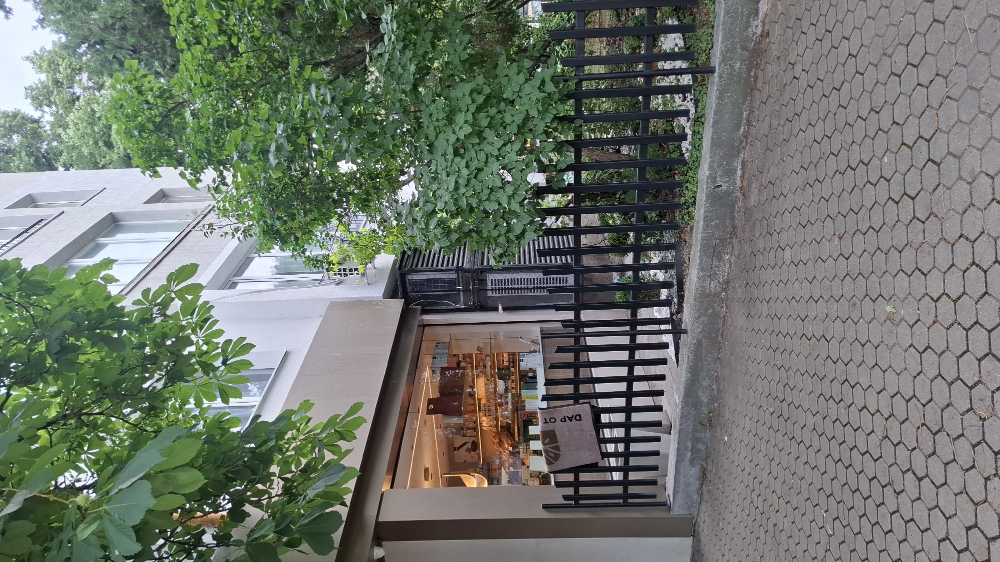 The image depicts the storefront of a bakery named "DAP OT" in Sofia, Bulgaria. The bakery is located on the ground floor of a multi-story building, with large glass windows showcasing the interior. Inside, shelves are stocked with various baked goods and products. A black metal fence encloses a small area in front of the bakery. Green trees and foliage are visible on the left and right sides of the building, as well as in the background, indicating an outdoor urban setting. The ground in front of the fence is paved with hexagonal grey stones. Two air conditioning units are mounted on the exterior wall of the building next to the bakery entrance. The lighting suggests it is daytime.