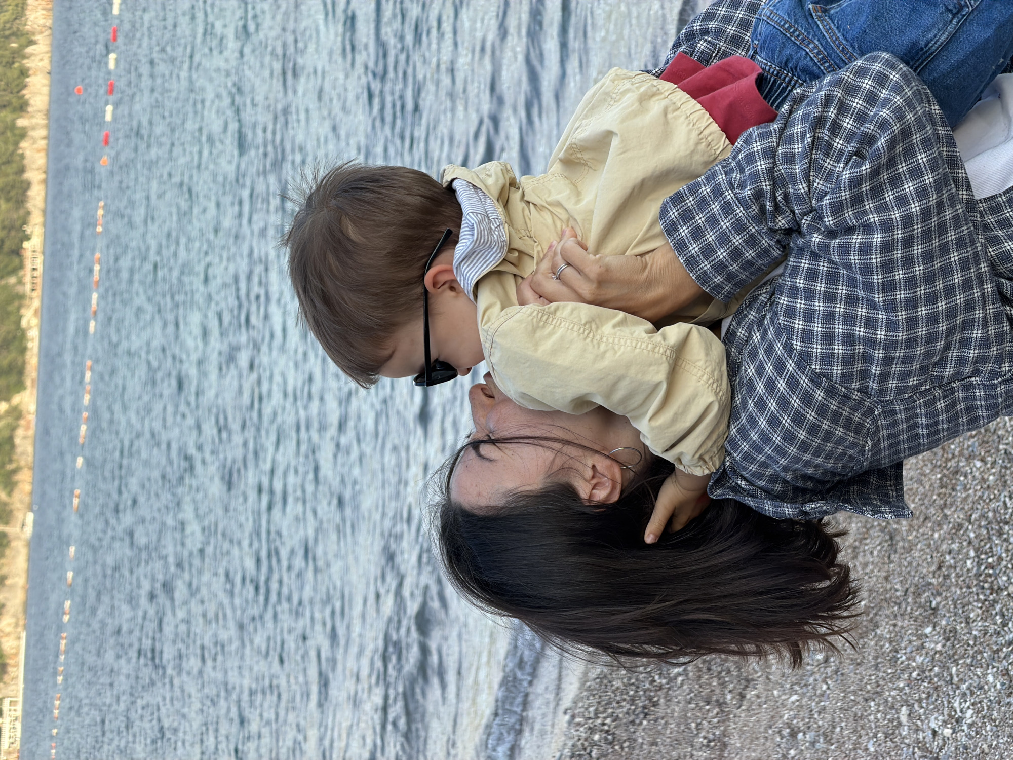 A woman and a child are lying on a pebble beach. The woman has dark hair and is wearing a blue and white plaid shirt, blue jeans, and hoop earrings. The child is wearing sunglasses and a tan jacket. They appear to be on a beach in Budva, Montenegro. A line of red and white buoys is visible in the background near the water's edge, and the water is calm. The image appears to be taken from above, looking down at them. The woman is lying down, and the child is lying on top of her, with their faces close together.