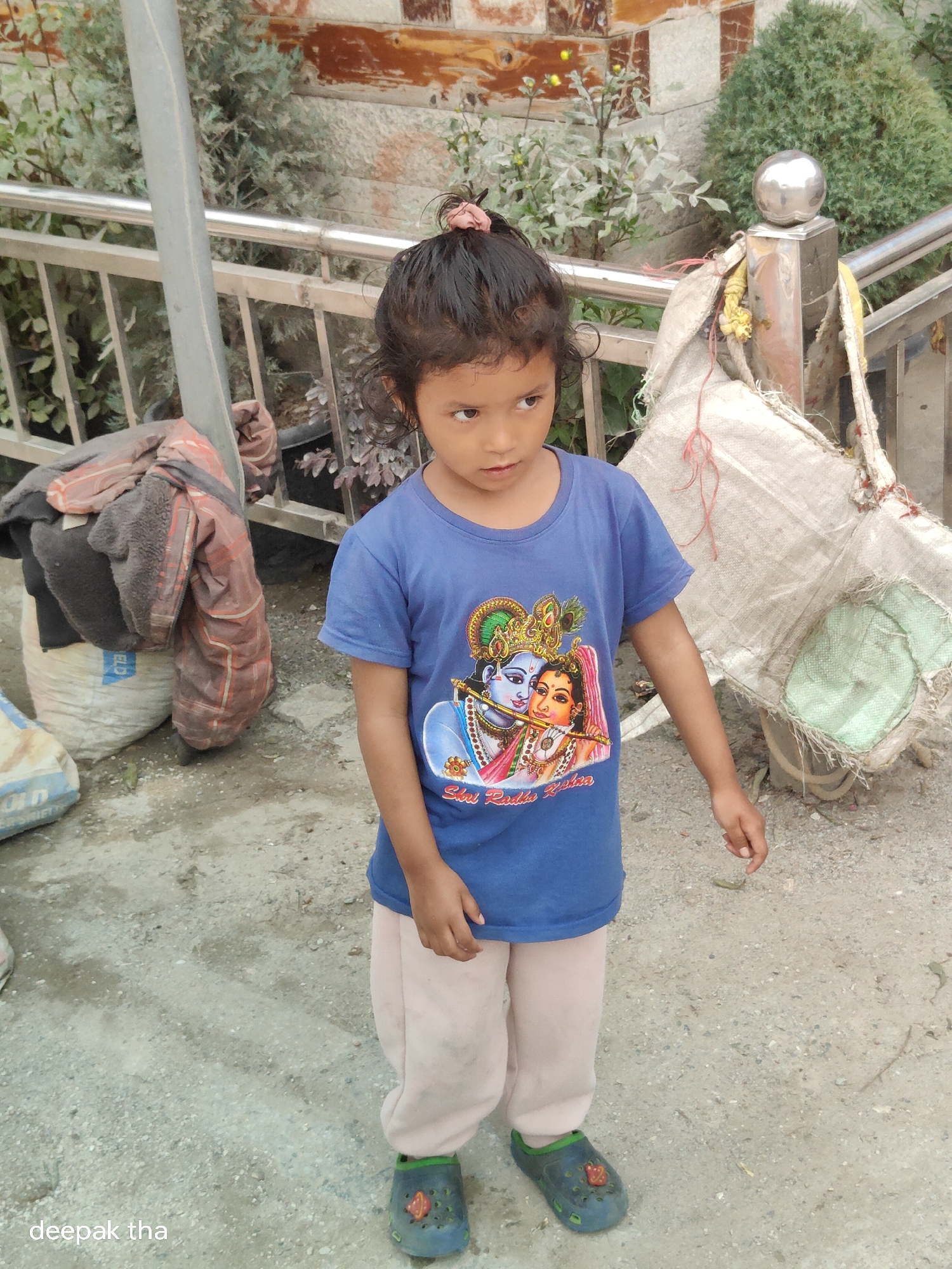 A young girl stands on a gravel path in Vashisht, India, wearing a blue t-shirt with an image of Radha and Krishna, and light-colored pants. She has dark, curly hair tied up with a pink scrunchie. Her eyes are looking to her right, and her right arm is bent at the elbow with her hand resting near her hip. She is wearing dark blue crocs with a red emblem. Behind her, to the left, are two large bags, one white and one beige, with blankets draped over them. A metal railing runs along the background, separating the path from some greenery and a stone and wood wall. A large, rolled-up canvas or burlap sack is tied to a metal post on the right side of the image. The overall scene suggests an outdoor setting possibly in a residential or public area in India.