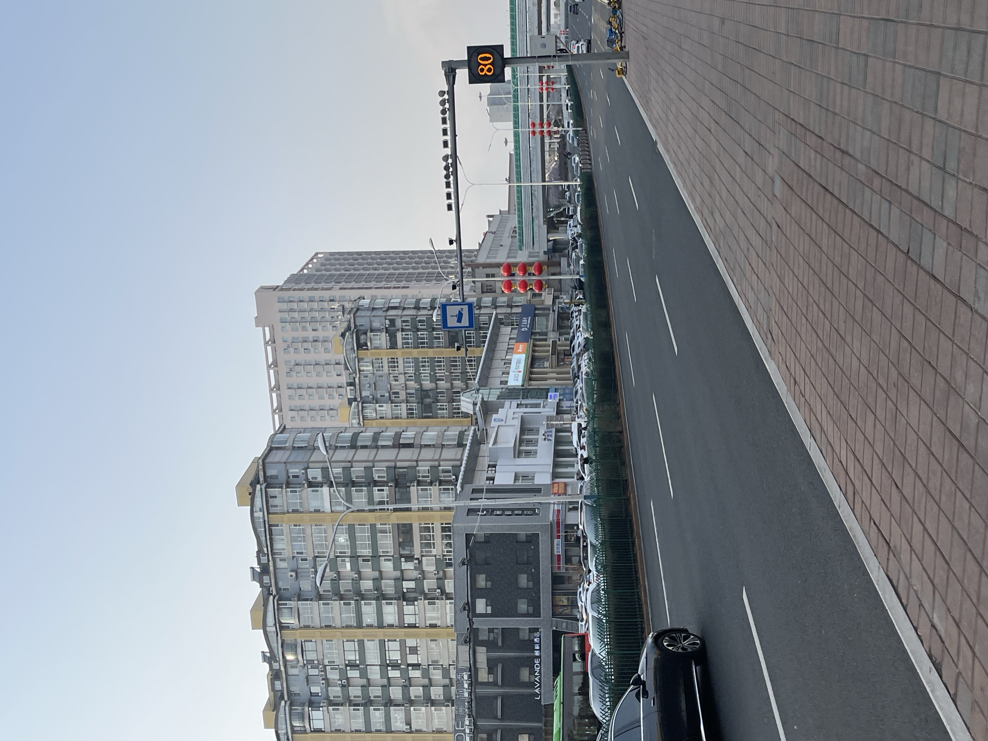 The image displays a clear daytime urban street scene in Changchun City, China. The perspective is from a slightly elevated point, looking down a multi-lane road that runs from the foreground into the distance. A wide, brick-paved sidewalk is visible on the right side of the road.
On the left, numerous modern high-rise buildings line the street, featuring a dense arrangement of windows and external air conditioning units. One building stands out with distinctive yellow-gold trim and an undulating or wavy architectural element on its facade.
Along the roadside, several vehicles are parked in a designated lane, and a black sedan is partially visible on the main road in the foreground. A row of yellow shared bicycles is parked on the sidewalk/shoulder area. Overhead, a prominent digital sign displays the number "80" in orange, likely indicating a speed limit or traffic speed. Below this gantry, a set of red traffic lights is visible. A blue rectangular road sign, featuring a white symbol resembling a vehicle and a station, is also present near the buildings.
Visible text includes the number "80" on the digital display and "LAVANDE 麗枫酒店" (Lavande Hotel) on the exterior of one of the buildings. The sky is clear and bright, suggesting fair weather and good visibility. The scene depicts a routine moment in a bustling city, with typical urban infrastructure and architecture.