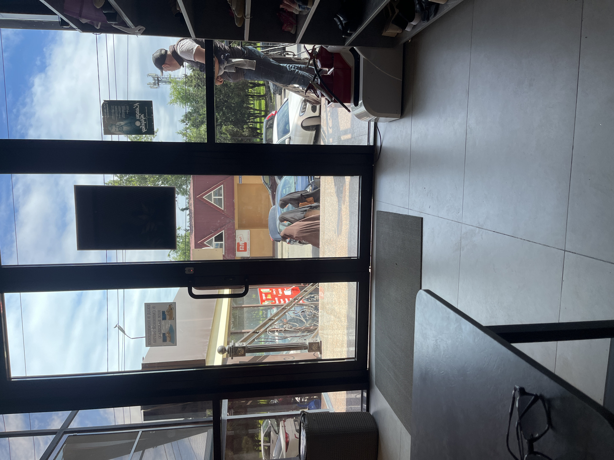 A view from inside a building looking out through a black-framed glass door. The door is slightly ajar, revealing a street scene in Kaspiysk, Russia. Outside, a man on a ladder is working near a white car. There is also other parked vehicles. Buildings with signage are visible across the street. The interior of the building features tiled flooring, shelving with shoes, and a table with eyeglasses. A gray rug lies near the door.