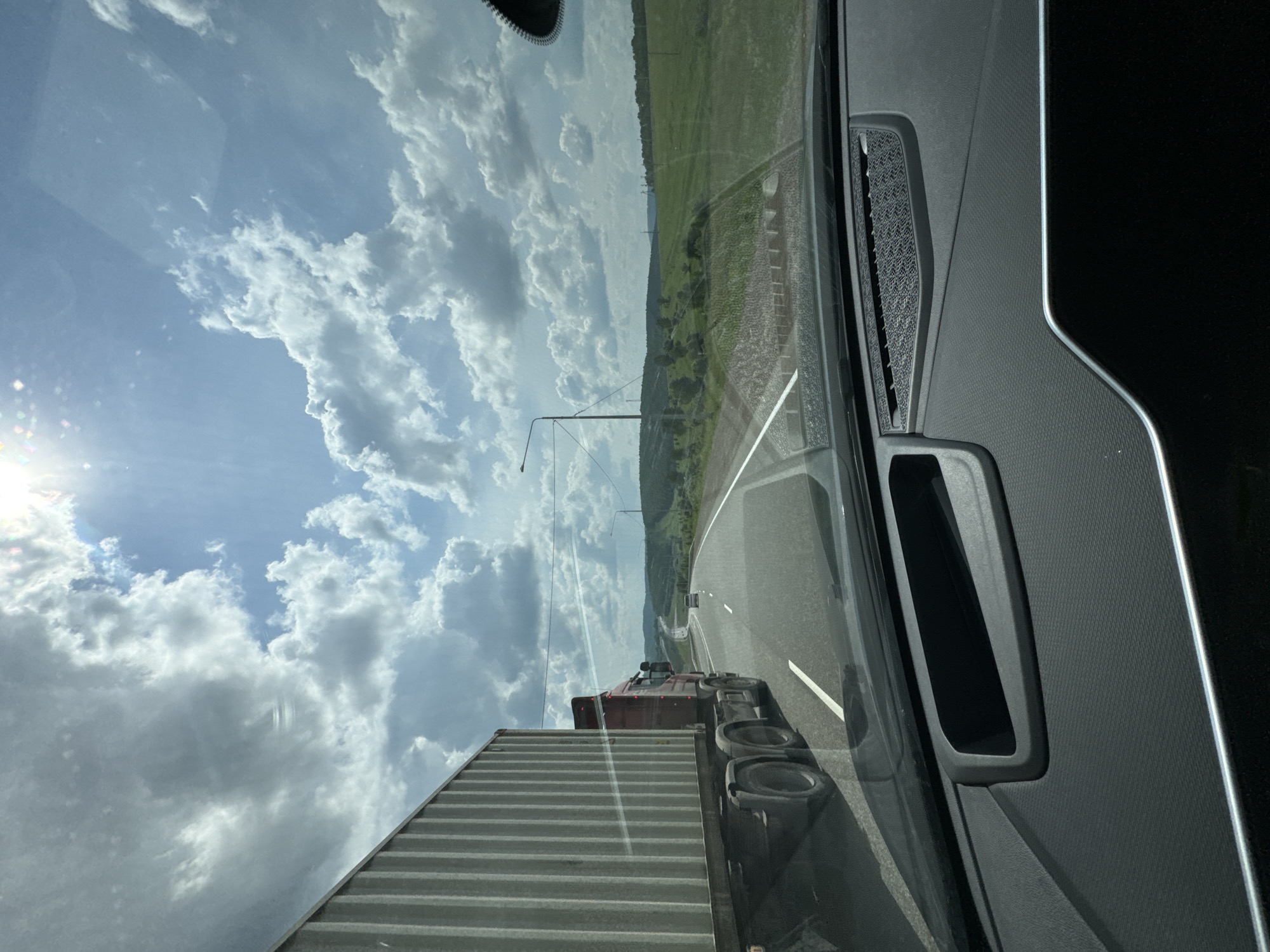 This is a forward-facing, eye-level, outdoor, daytime, photographic image taken from inside a vehicle. The perspective is from the driver's seat, looking through the windshield. The vehicle is traveling on a multi-lane asphalt road. The road is bordered by green fields and rolling hills under a partly cloudy sky. The sun is visible in the upper portion of the frame, casting a glare on the windshield. A large red semi-truck with a white cargo container is in the left lane, traveling in the same direction as the camera vehicle. There are other vehicles visible in the distance on the road. The interior of the car's dashboard is visible in the foreground. The location is identified as Voznesenka, Russia.