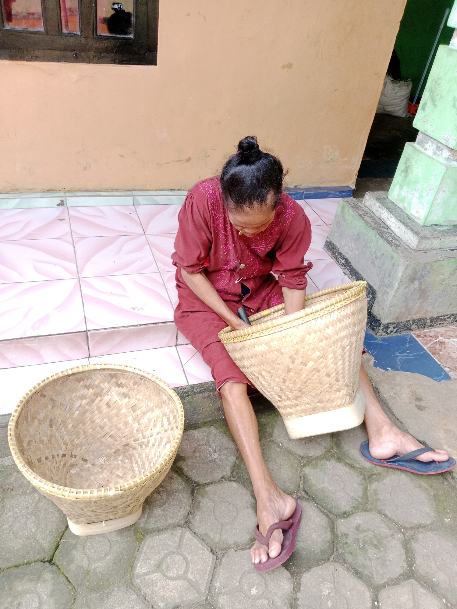 An elderly woman with her hair in a bun, wearing a dark red traditional Indonesian garment, is sitting on the ground in front of a light orange wall. She is working on a woven bamboo basket, holding it with her hands and looking down at it. To her left, another similar woven bamboo basket sits on the ground. The ground is paved with hexagonal concrete tiles and some rectangular pink tiles in the foreground. There is a window with dark brown frames on the upper left of the wall. To the right, a green structure made of stacked concrete blocks is visible. A white sack is leaning against the wall in the background. The scene appears to be outdoors, possibly in a residential area or courtyard in Parage, Indonesia.