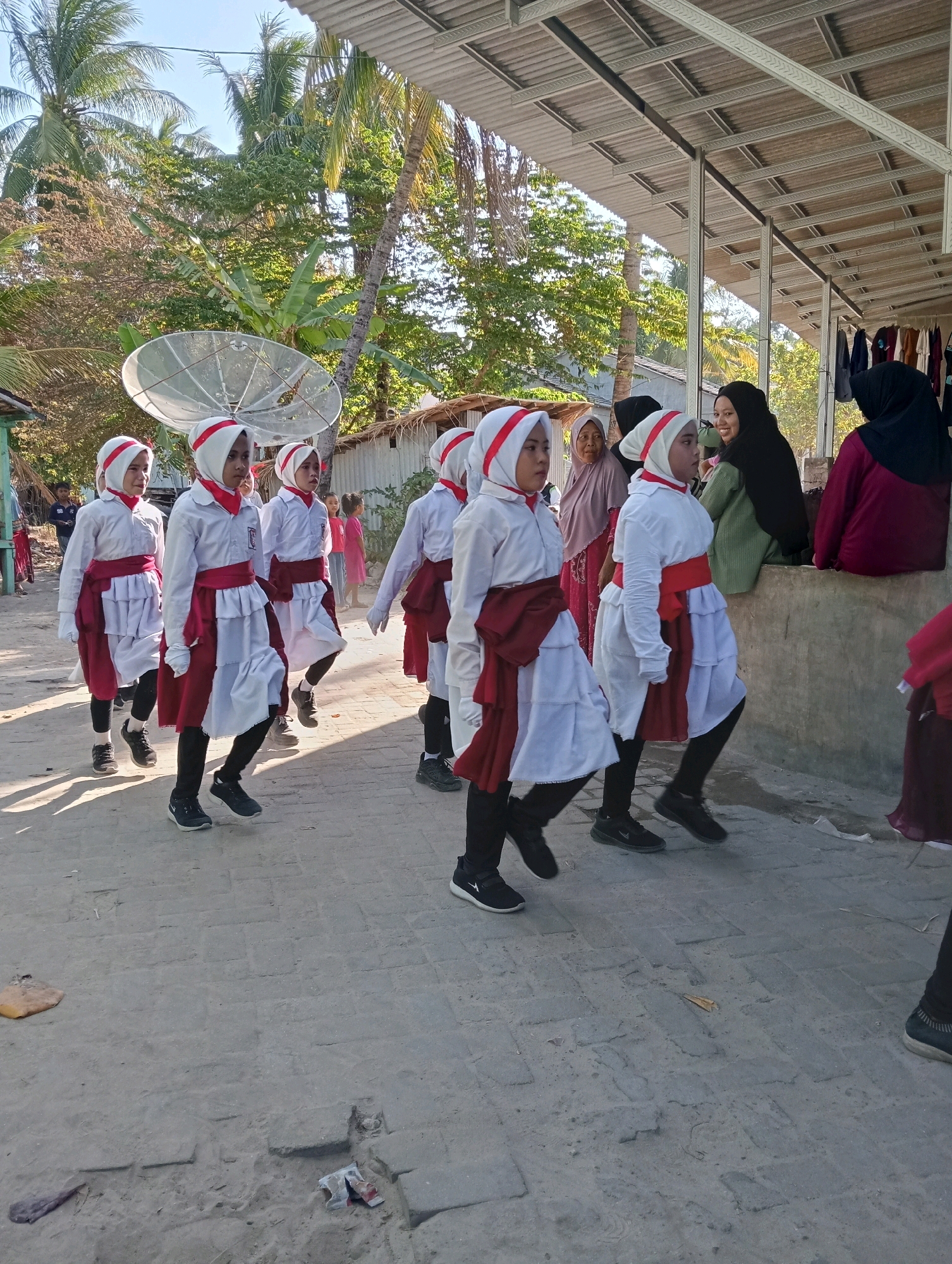 In Pagerungan Kecil, Indonesia, a group of young girls dressed in white uniforms with red sashes and headscarves are marching in formation on a paved pathway. Their uniforms consist of white shirts, layered white skirts with red underskirts, and black leggings. They are walking towards the right of the frame. Behind them, several adults are observing, including a woman in a green hijab and another in a dark maroon hijab, who is seated. The environment features a corrugated metal roof structure on the right, with clothing hanging on display. Tall palm trees and other foliage are visible in the background, along with a satellite dish. The ground is a mix of paved bricks and dirt. The lighting suggests it is daytime, with sunlight casting shadows.