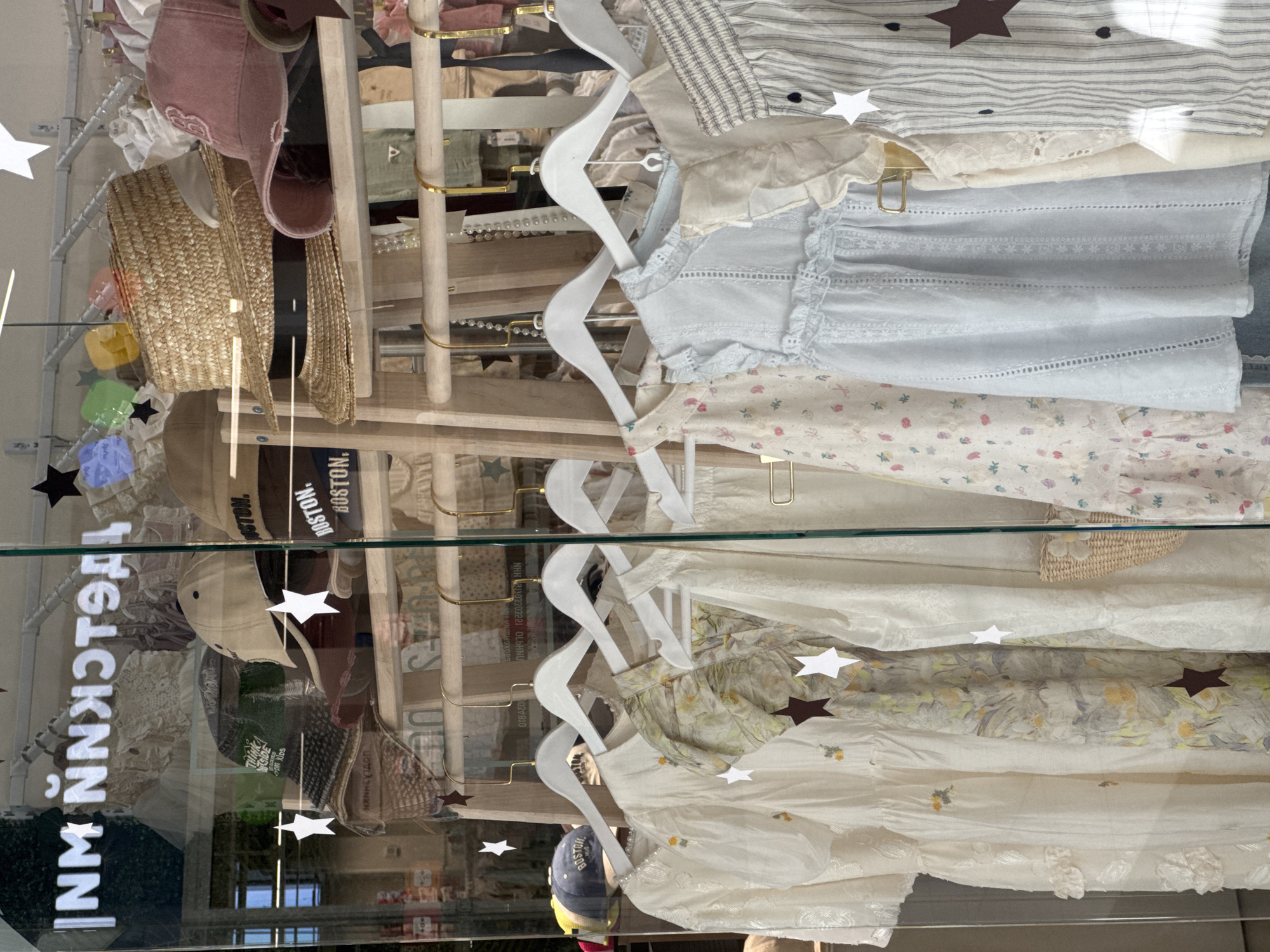 The image shows a display window of a clothing store in Mikhaylovsk, Russia. The window displays a variety of children's clothing, primarily dresses and tops, in light colors and floral patterns. There are also several hats, including straw hats and baseball caps, visible on shelves behind the clothing. Decorative stars are placed on the glass and on some of the garments. The store's name, "NM ŇNKGT9A", is reflected on the glass, written in white block letters. The lighting in the image is bright, suggesting it is daytime.