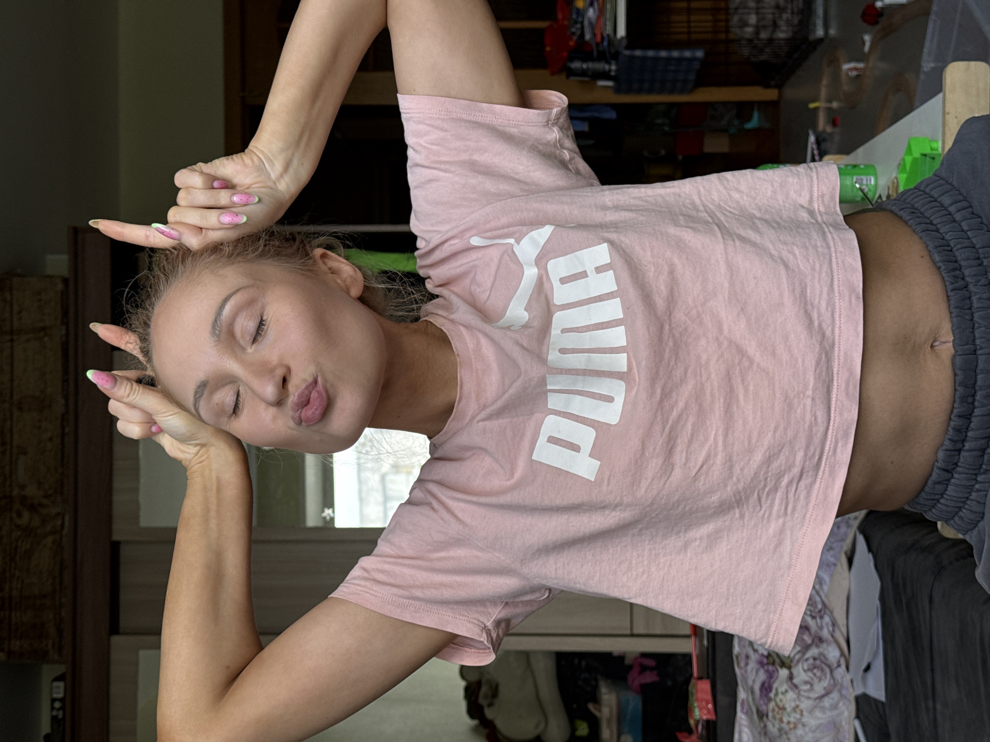 A fair-skinned female with blonde hair and watermelon-themed nail art poses indoors, making a playful gesture with her hands near her head. She is wearing a light pink Puma t-shirt and grey athletic shorts. The background appears to be a cluttered room with furniture and various items on shelves. The location is specified as the city of Chaikovsky, Russia.