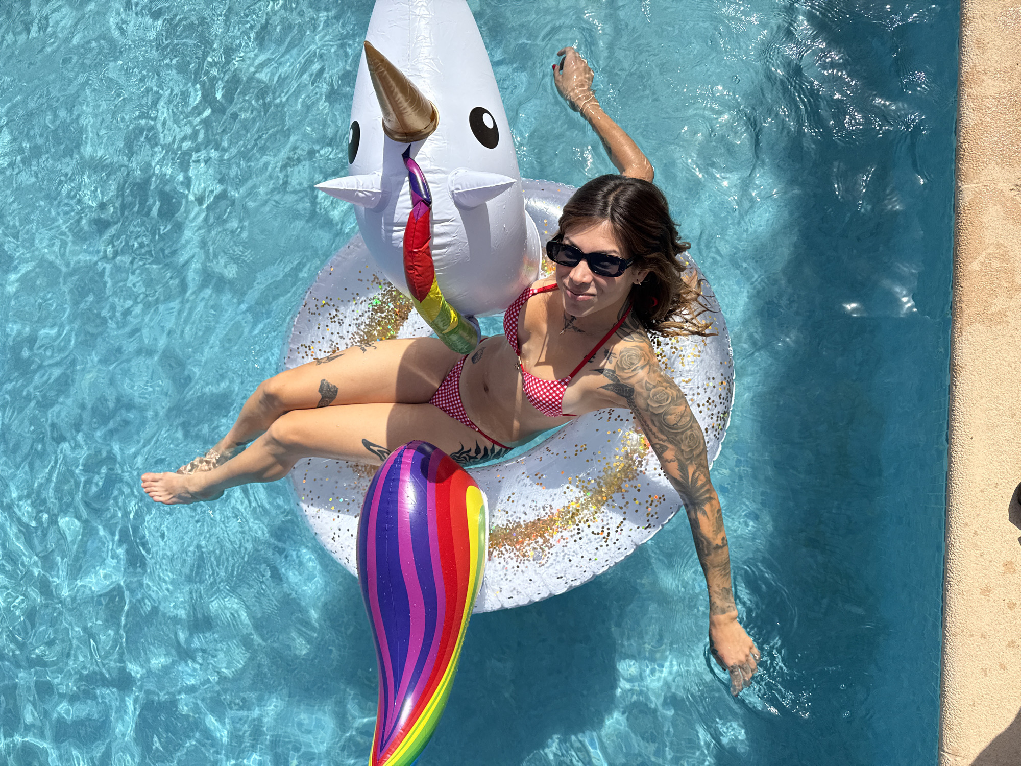 A woman with extensive tattoos on her arms and legs lies on her back on an inflatable unicorn float in a swimming pool. She is wearing a red and white checkered bikini and black sunglasses. The pool is filled with clear blue water. The unicorn float is white with a golden horn and black eyes. A rainbow-colored inflatable is attached to the front of the unicorn. The scene is lit by bright sunlight, casting shadows on the water. The location is the city of Sant Vicenç de Montalt, Spain.
