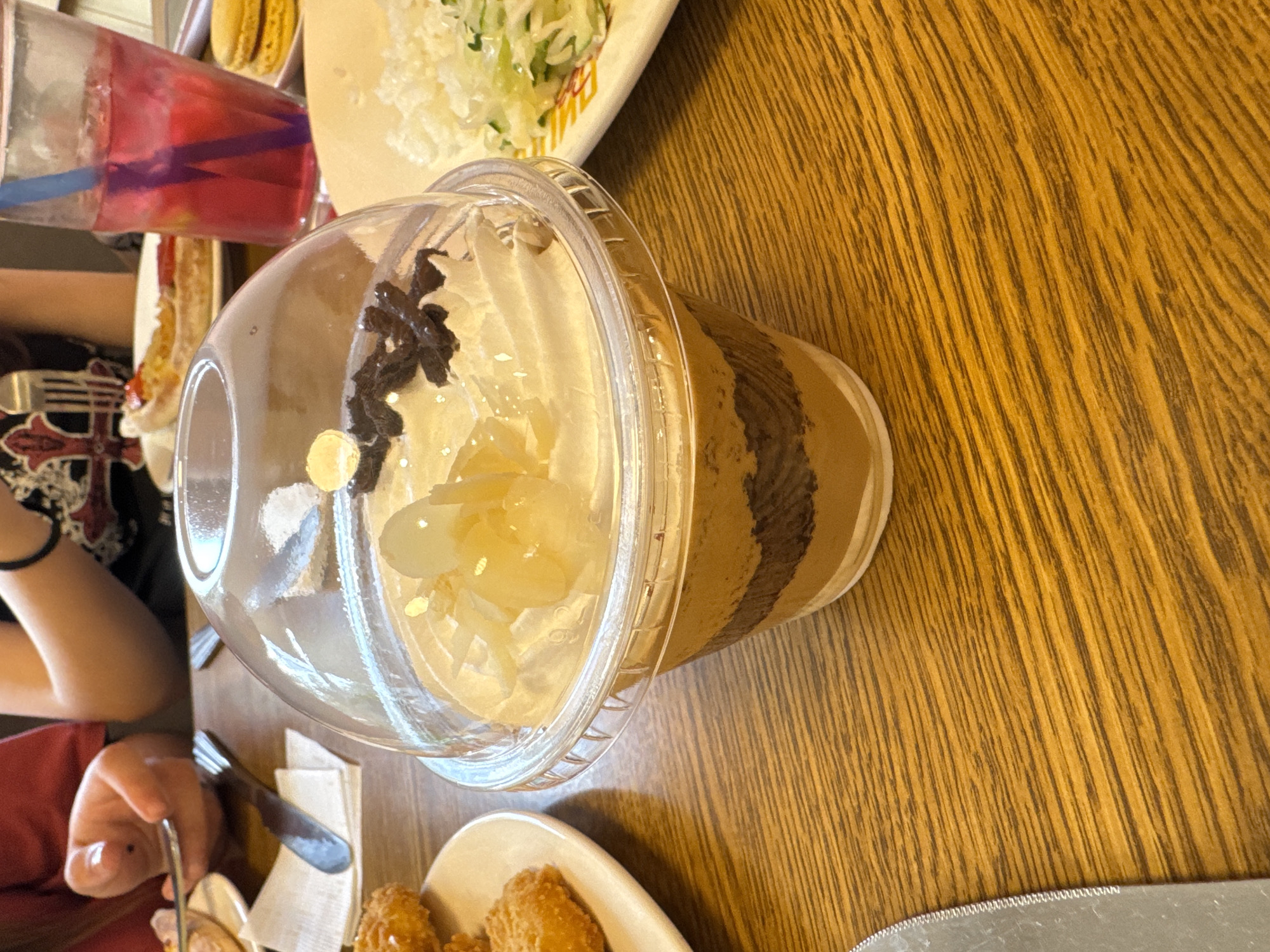 The image shows a close-up of a dessert in a plastic cup with a domed lid, placed on a wooden table. The dessert appears to be layered with a light brown substance and a darker brown substance, topped with whipped cream, chocolate shavings, and what looks like pieces of candied fruit. To the left of the dessert, a fork is resting on a white napkin, and a person's hand is visible, holding another fork. In the background, a glass of pink beverage with a blue straw is visible, along with a plate of macarons and another plate with a portion of what seems to be a strawberry tart. A person wearing a black and red patterned shirt is also partially visible in the background, and another person in a red shirt is to the far left. The lighting suggests an indoor setting, possibly a cafe or restaurant. The city of Oskemen, Kazakhstan, is not depicted in this image; the image is a close-up of a table setting with food and drinks.