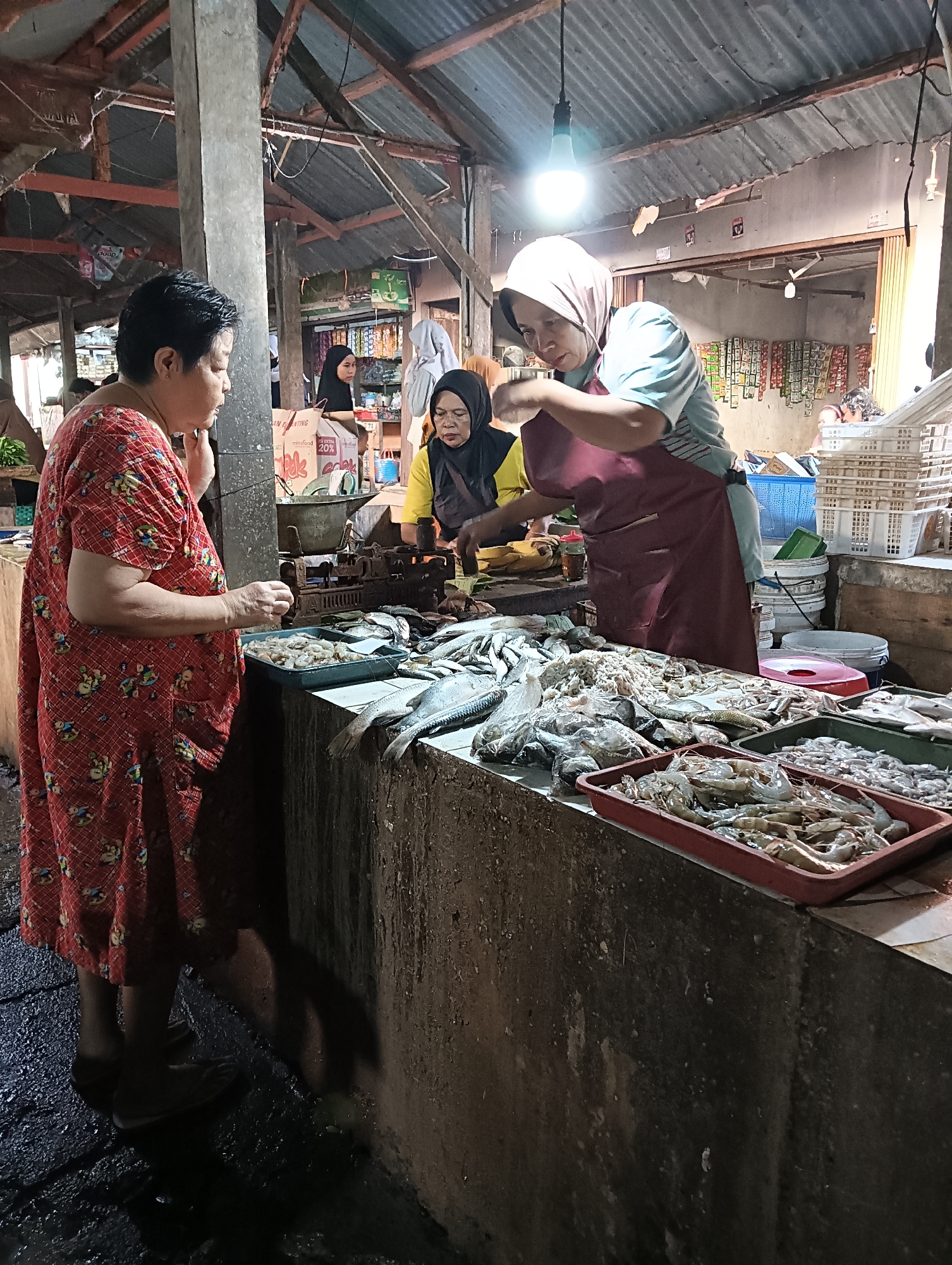 In a dimly lit outdoor market stall in Cluwak, Indonesia, a woman wearing a red patterned dress stands at a counter filled with various types of fish and shrimp. She is facing a woman in a light blue shirt and burgundy apron, who is tending to the seafood. The woman in the apron is looking down at the counter and appears to be serving or arranging the seafood. Another woman in a yellow shirt and headscarf is visible behind the counter, also engaged in market activities. The stall is constructed with a corrugated metal roof and wooden beams, and the counter is made of concrete. Various containers and scales are present on the counter, along with stacks of plastic crates in the background. The ground is wet and appears to be made of rough pavement.