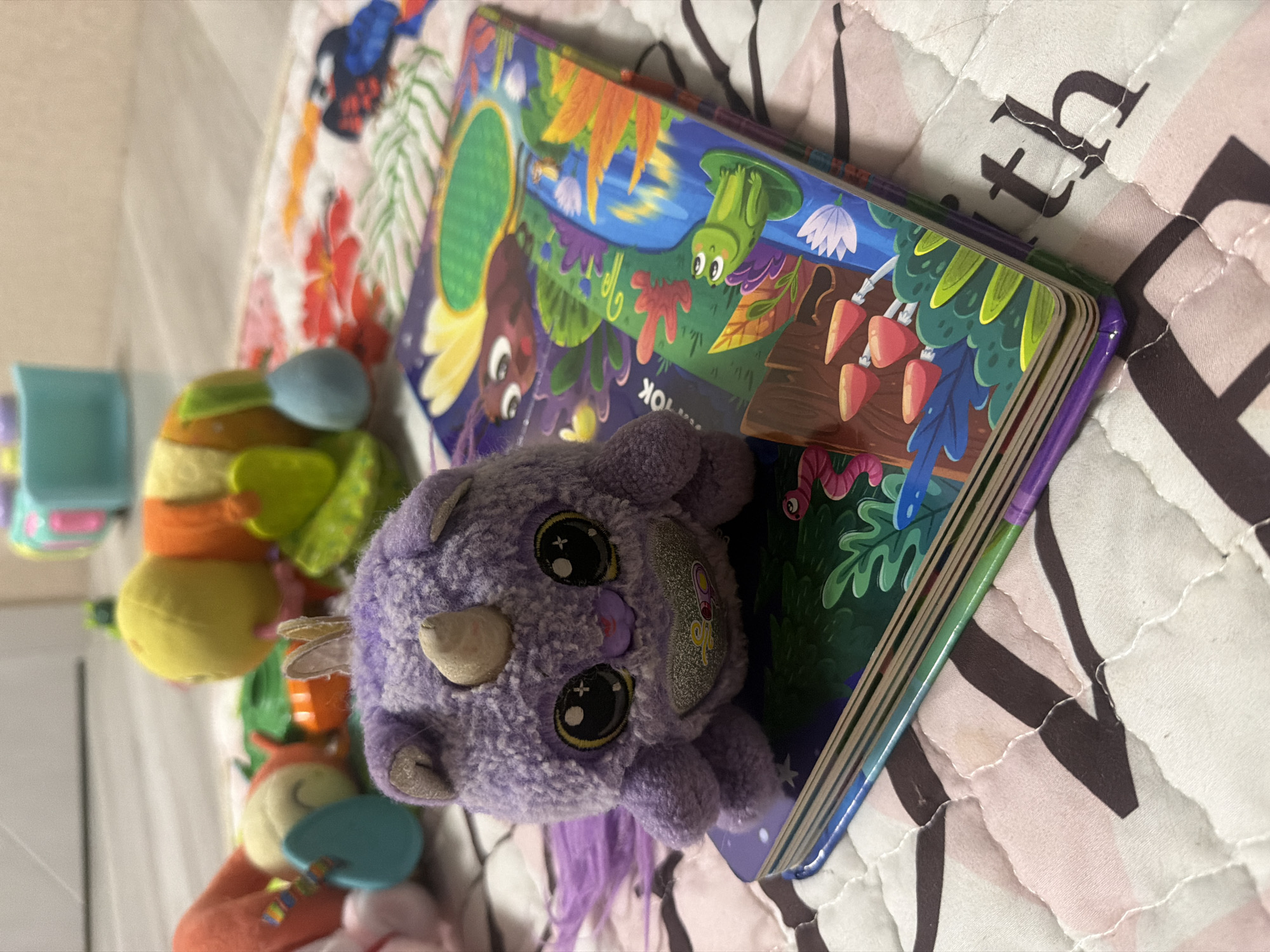 The image shows a purple plush unicorn toy with large black eyes and a white horn, sitting on top of an open children's book. The book has a colorful illustration on its cover, featuring a frog, a butterfly, and various plants. The scene appears to be indoors, possibly on a quilted playmat with a floral pattern. Several other children's toys are visible in the background, including a caterpillar-shaped toy and a blue-green toy with stacked rings. The lighting suggests a casual indoor setting. There is no information in the image to determine its location in Rostov-on-Don, Russia.