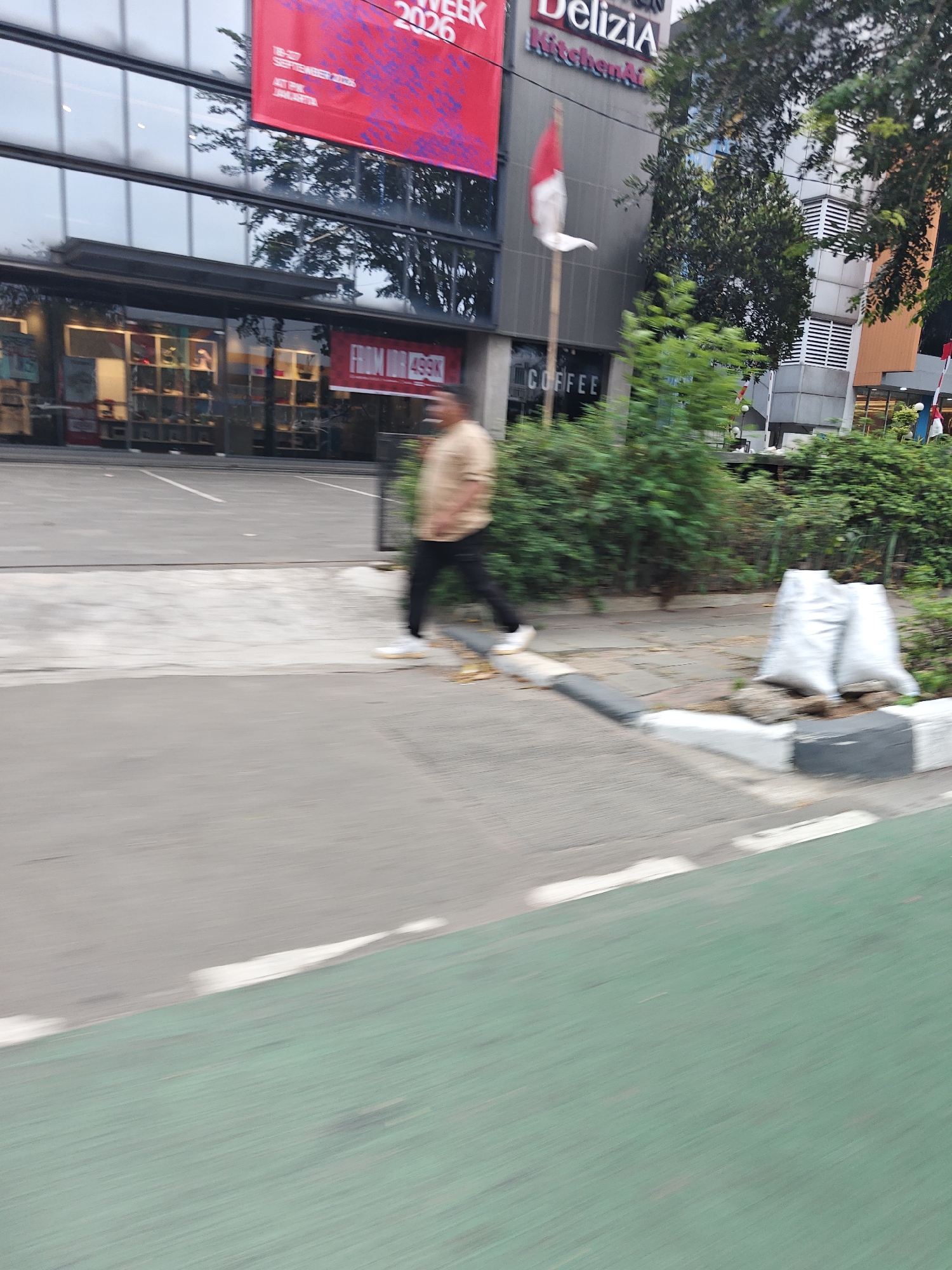 A horizontal, slightly blurred, eye-level outdoor shot depicts a man in a tan shirt and dark pants walking across a paved area with a green bike lane in the foreground. The man is in motion, with his arms slightly extended and his legs in stride. Behind him, a modern building with large glass windows and a red banner advertising "WEEK 2026" is visible. The building also has signage for "Delizia Kitchenware" and "COFFEE." A few trees and bushes are scattered around the scene, and two white sacks are placed near the curb on the right side of the image. The overall lighting suggests it is daytime.