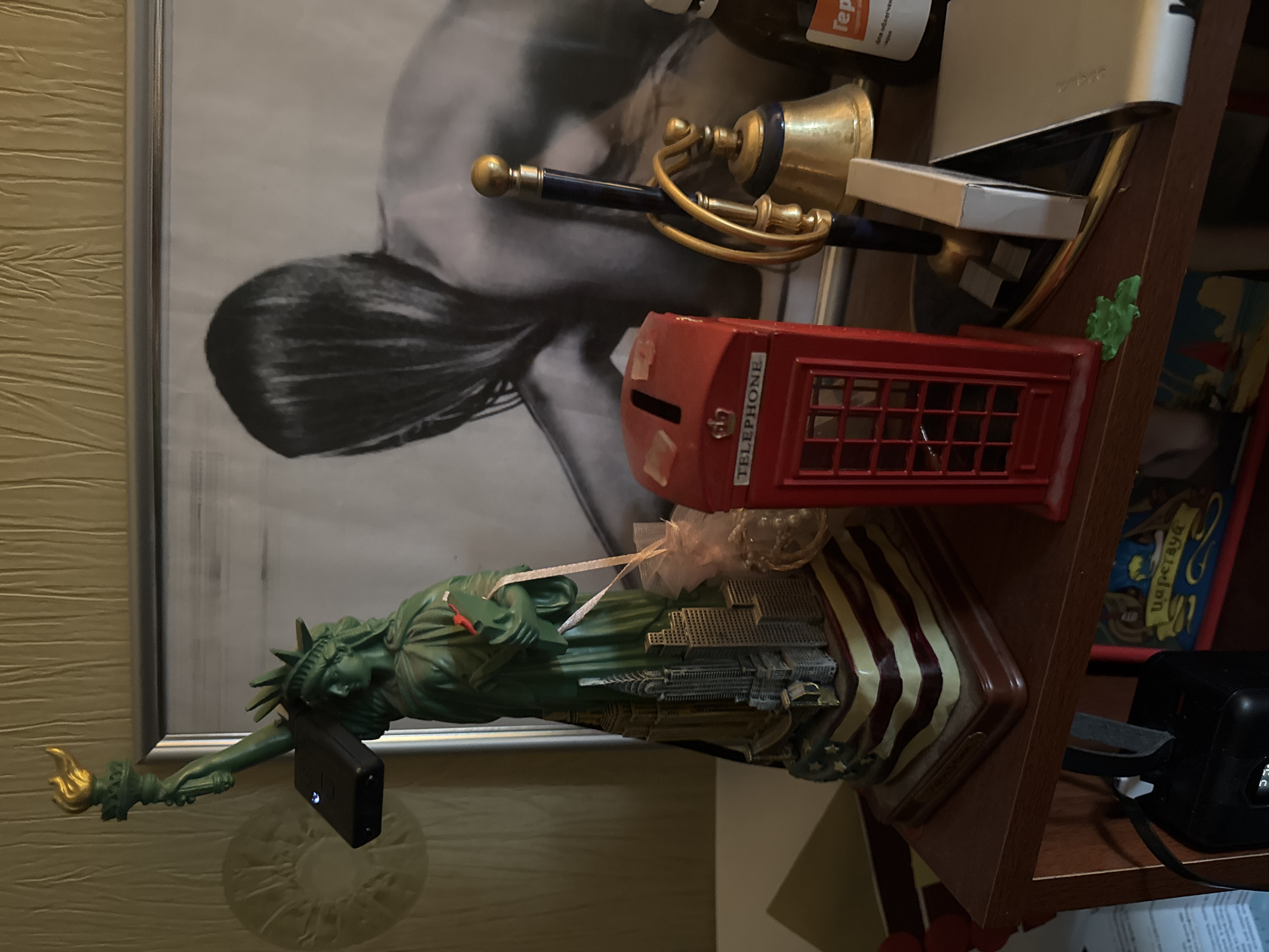 The image depicts a shelf displaying various decorative objects and a framed picture. Prominently featured is a statue of the Statue of Liberty, detailed with buildings at its base and a base designed like the American flag. Attached to the statue's torch arm is a black electronic device with a blue light. To the right of the statue is a miniature red British telephone booth coin bank. Further to the right, a brass bell with a blue-accented handle stands next to a blue and gold desk bell. Behind these objects is a framed black and white photograph of a person with long dark hair. The shelf is made of dark wood, and a portion of a yellow, textured wall is visible above it. A bottle with Russian text is partially visible on the far right. The scene appears to be an indoor display, possibly in a residential setting, with no discernible connection to Sertolovo, Russia, based on visual evidence alone.