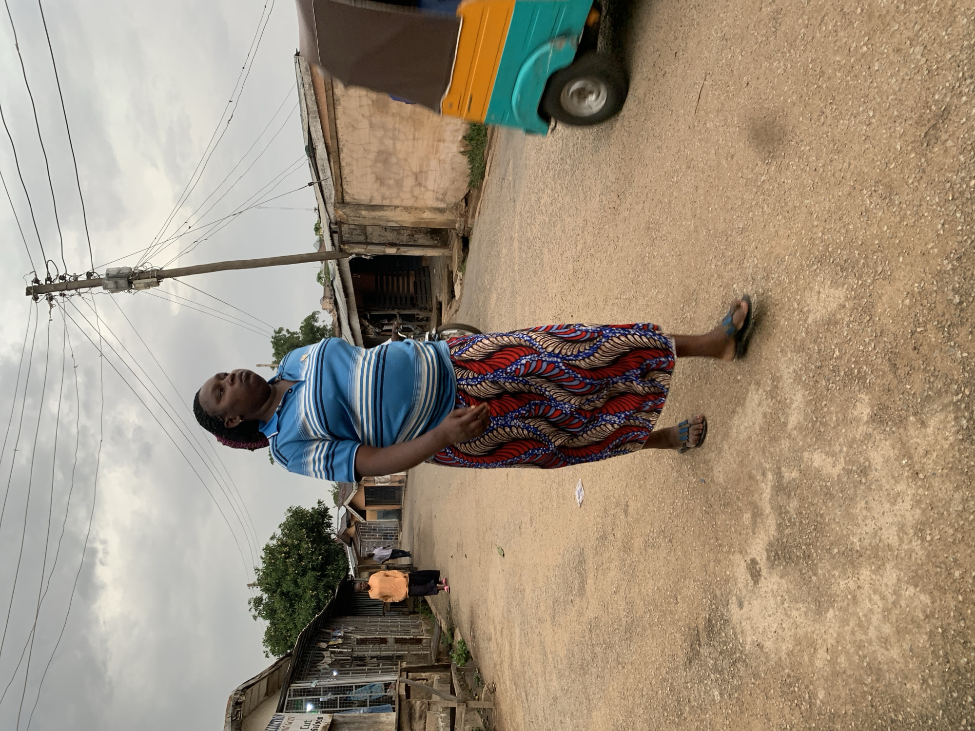 The image shows a woman standing on a paved road in Jos, Nigeria. She is wearing a blue striped shirt and a long, patterned skirt with red, blue, and white colors. She is looking up. In the background, there are buildings, utility poles with numerous wires, and an auto rickshaw. The sky is overcast, suggesting it is daytime. Another person is further down the road.