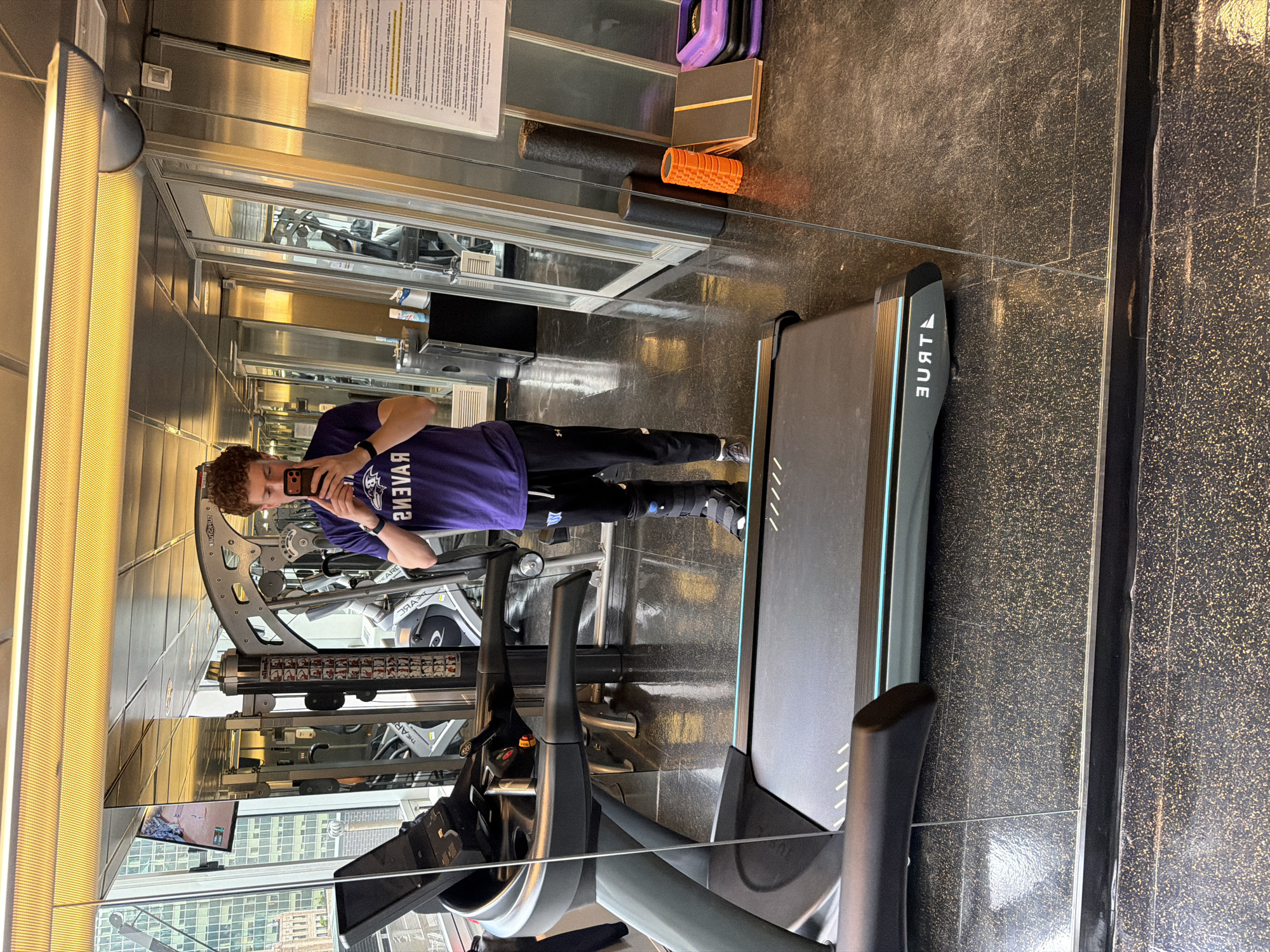 The image depicts a person in a gym, visible in a mirrored wall. The person is taking a selfie with their phone. They are wearing a purple t-shirt with the word "Ravens" printed backwards on it, black pants, and a medical boot on their right foot. They stand next to a "True" brand treadmill.
The gym appears to be located in a high-rise building in New York City, visible through a large window. Exercise equipment is seen throughout the gym, including weight machines. The gym also contains foam rollers, and platforms, likely for stretching and workout routines. A sign is affixed to the wall near the top of the mirror. The gym floor is tiled.