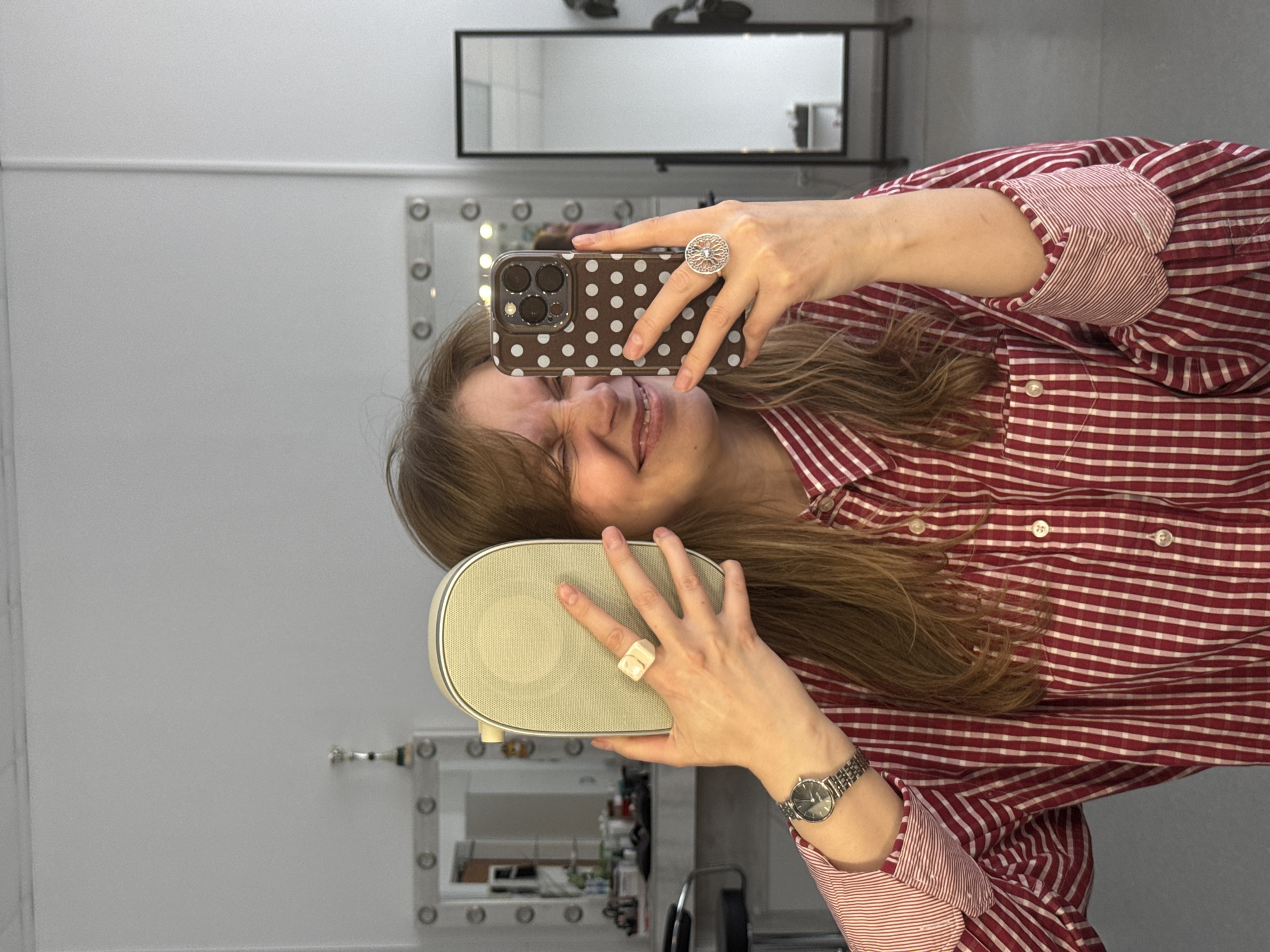 This sideways mirror selfie features a young woman smiling broadly, her eyes squinted in joyful amusement. She is positioned horizontally across the frame. The indoor setting, likely a dressing room or studio in Izhevsk, Russia, has white or light grey walls. In the background, a long, rectangular mirror reflects the ceiling, and below it, a smaller vanity mirror with exposed bulbs reflects various unseen items.
The woman has long, light brown hair falling downwards (relative to her body). She wears a red and white checkered long-sleeved shirt with rolled-up cuffs that reveal a contrasting red and white striped pattern. In her right hand, extended towards the left, she holds a smartphone with a dark brown case adorned with white polka dots, used to capture the image. A large, intricate silver-colored ring with an openwork design is on her right middle finger.
In her left hand, held near her face, she grasps a light beige, rectangular portable speaker or similar electronic device, which has a textured, perforated top surface. On her left ring finger, she wears a chunky, irregularly shaped ring with white and beige swirls, and a silver wristwatch with a dark face is on her left wrist. No discernible text is visible on any objects.