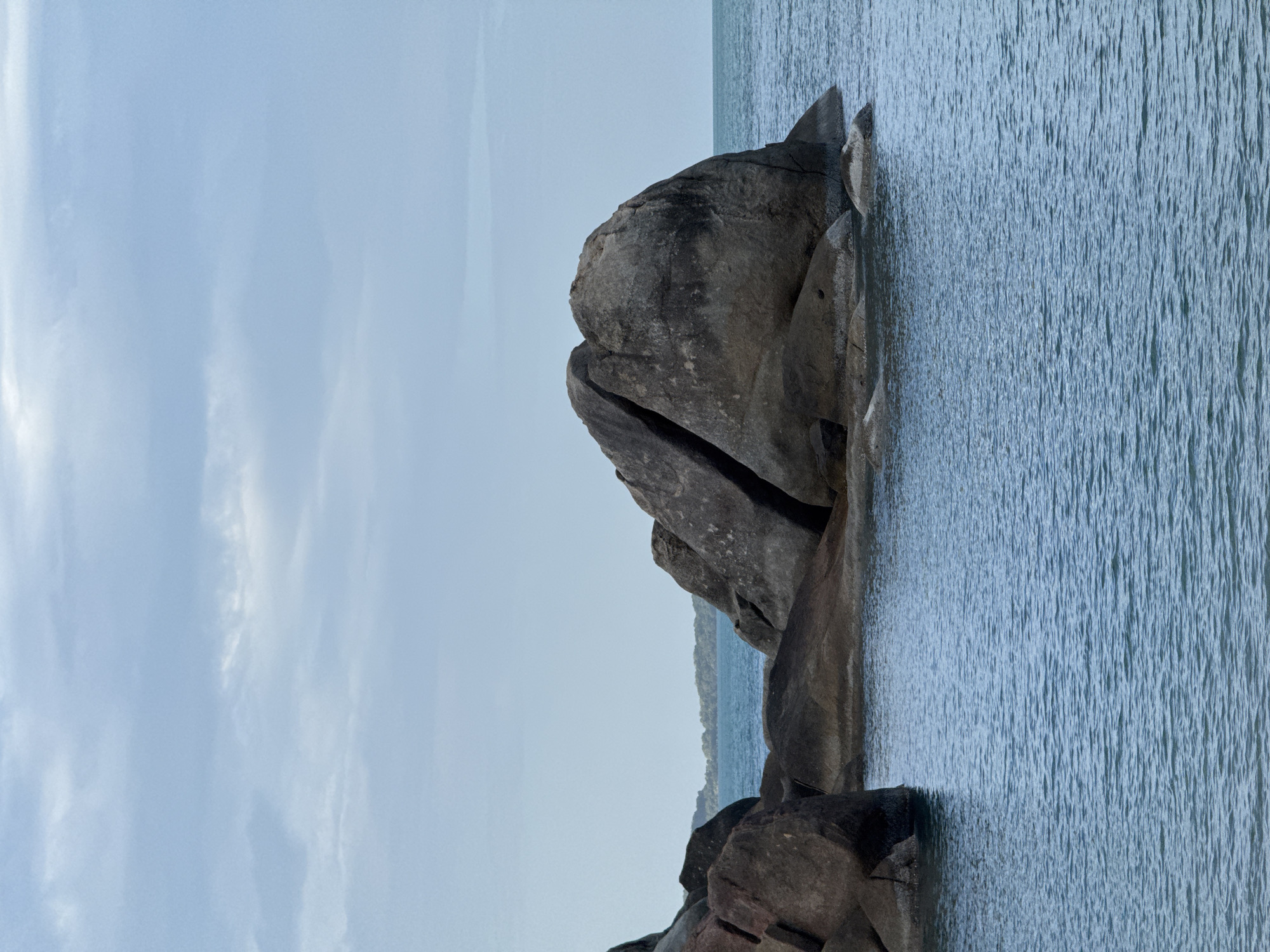 This image depicts a body of water with large boulders emerging from it. The water is calm with gentle ripples. The boulders are dark gray and appear to be of varying sizes and shapes, with some showing distinct fractures or crevices. In the background, a lush green shoreline with trees is visible, suggesting a coastal environment. The sky above is overcast with light blue and gray clouds. This scene is characteristic of the beaches in Ko Samui District, Thailand, known for its distinctive granite rock formations. There are no people or animals visible in the image.