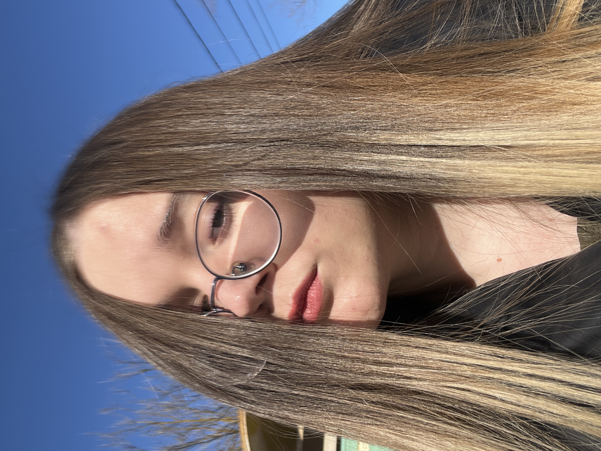 A close-up portrait depicts an individual with long, straight, light brown and blonde hair, which partially obscures the left side of the face and extends across the chest. The subject wears round-framed eyeglasses; the frame casts a prominent shadow across the right eye and upper cheek. The lips are tinted with a reddish hue. A small pink lesion is visible on the forehead. The individual is dressed in a dark-colored upper garment, possibly black, with a minimal visible portion of an olive-green fabric at the bottom right. The background features a clear, vivid blue sky. On the left, bare tree branches are visible, along with a partial view of a light-colored structure exhibiting turquoise or mint green elements. Faint utility lines are present in the upper right quadrant of the sky. The scene is illuminated by strong, direct sunlight, creating distinct highlights and shadows. The image was captured outdoors in the city of Большевик, Russia.