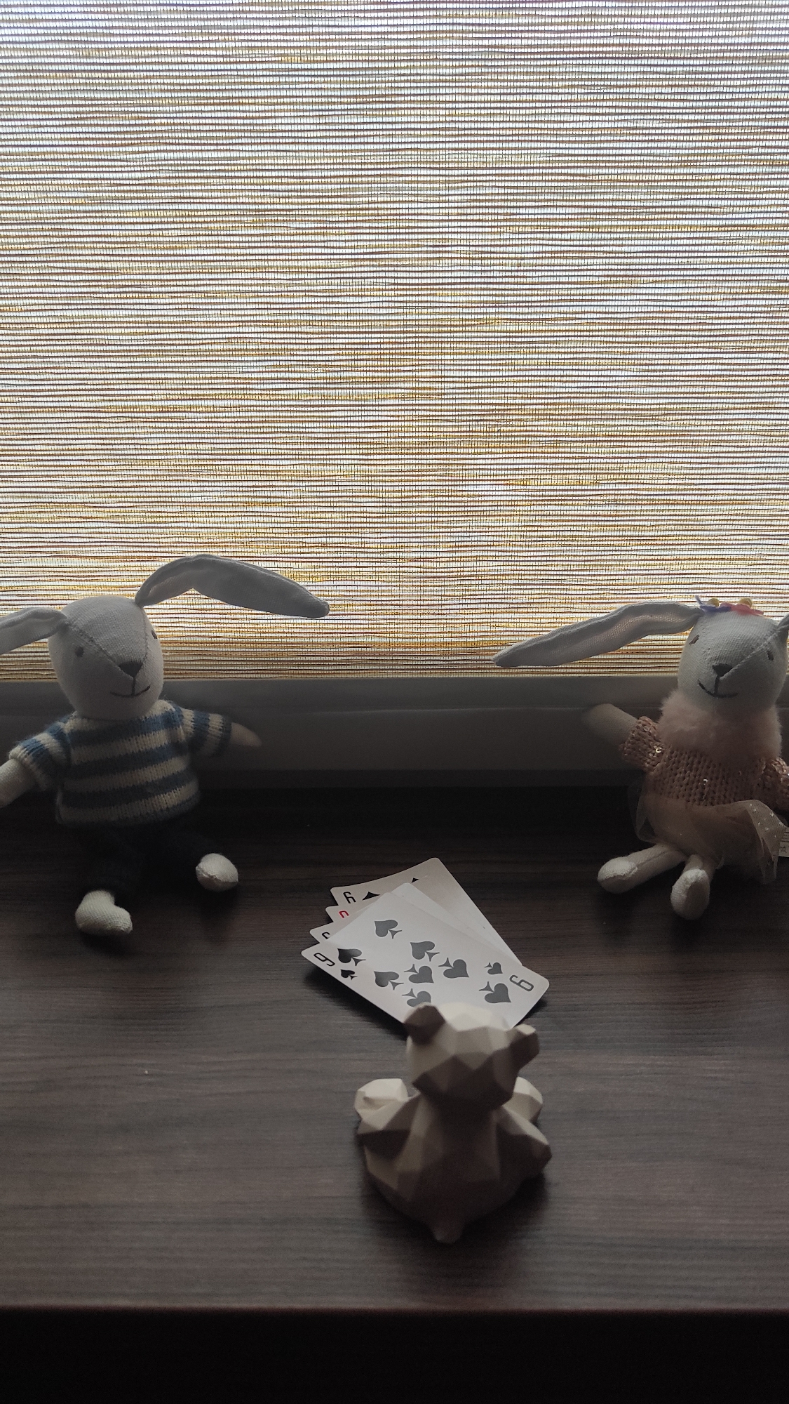 Two stuffed rabbit toys, a small geometric bear figurine, and a scattering of playing cards are arranged on a dark wooden surface. One rabbit wears a blue and white striped shirt, while the other wears a pink outfit. The cards, showing the six of spades, are partially obscured. A textured, light-colored window blind provides the backdrop. The location is Chaikovsky, Russia.