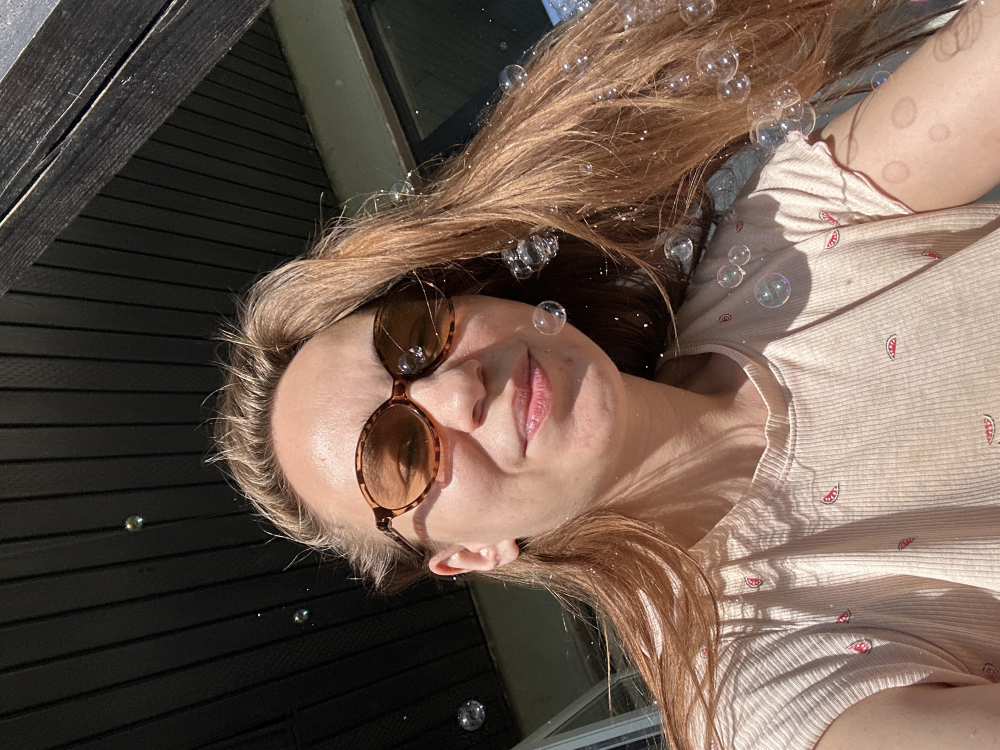 A woman with long brown hair and sunglasses is seen in a close-up, rotated perspective. Bubbles are floating around her head and hair. She is wearing a light pink top with watermelon patterns. The background includes a dark wooden structure. There is no indication of the city of Kharnavy, Russia.