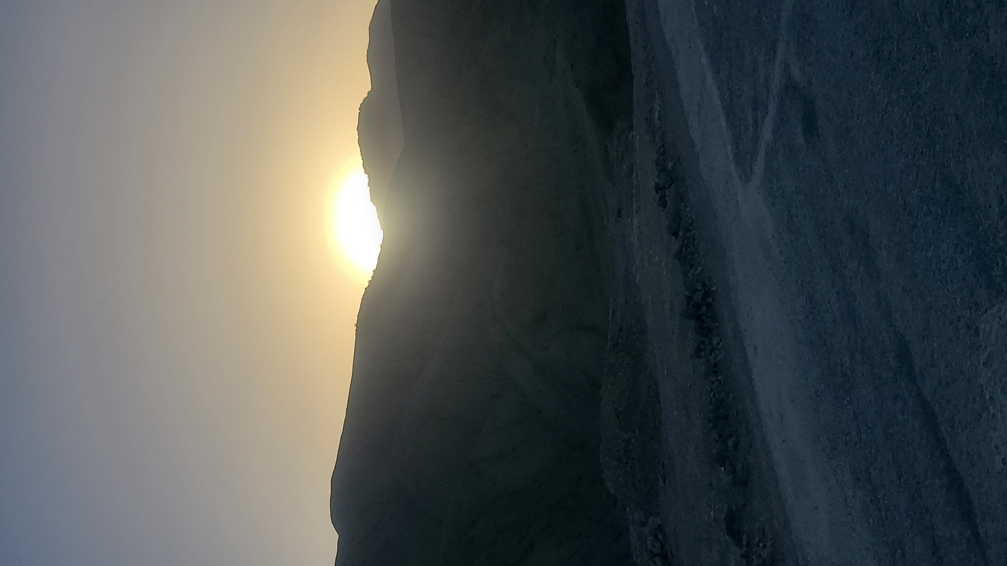A photograph taken in Ziarat, Pakistan, presents a stark contrast between the bright sun and the silhouette of a dark mountain. The sun, a radiant orb, peeks over the mountain's ridge, casting a golden glow on the surrounding sky. The mountain itself is a dark, undefined mass, with its texture and details obscured by the backlighting. To the right of the mountain, the terrain appears rough and uneven, possibly a path or a steep hillside. The overall impression is one of rugged beauty, with the stark contrast creating a dramatic and somewhat otherworldly effect.