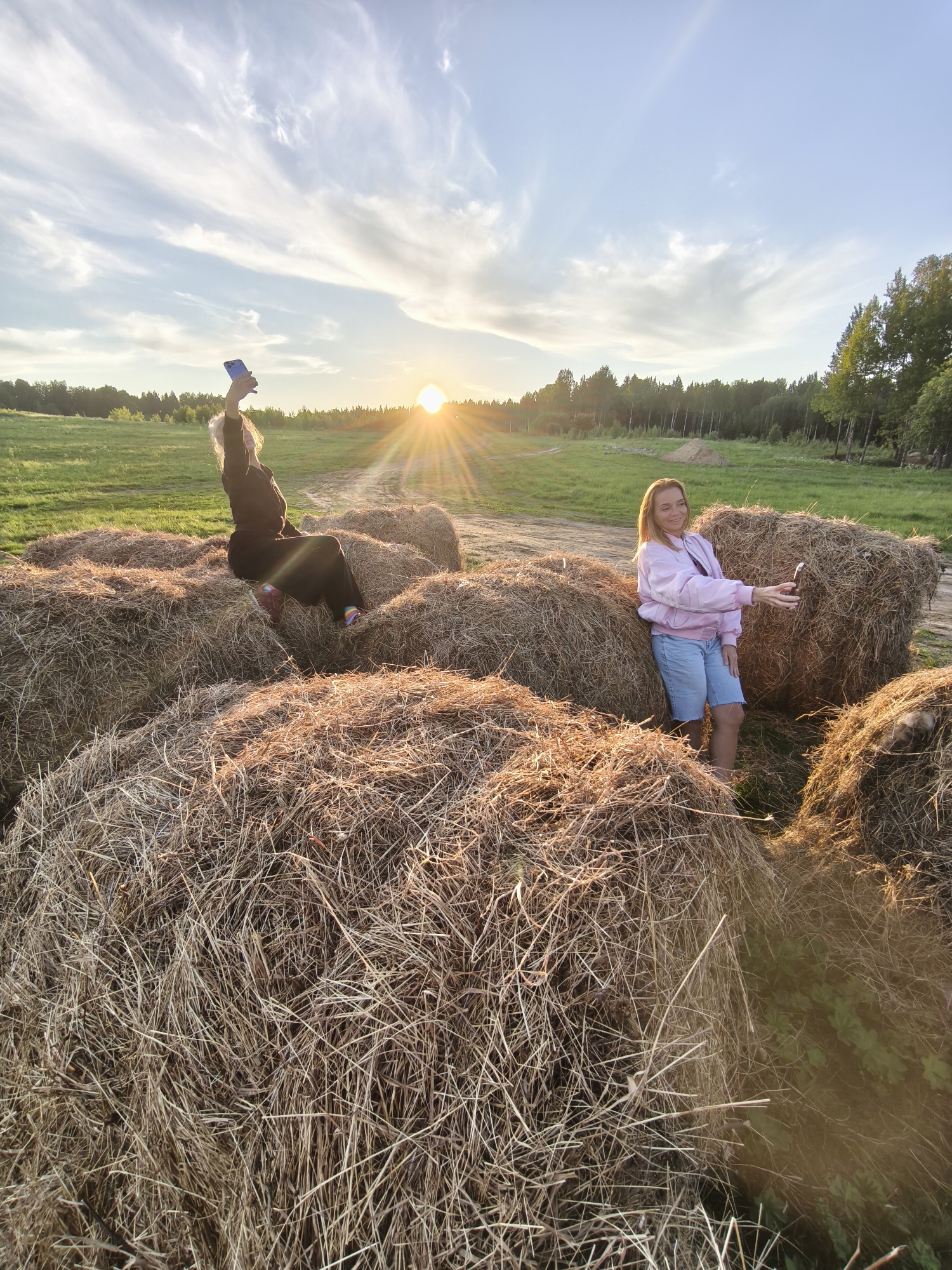 The image captures a serene rural scene at sunset in Стеклянный, Russia, featuring two women amidst several large, round hay bales. The sun, positioned low on the horizon to the left, casts warm, golden light and prominent lens flares across the field, signaling the golden hour. The sky above is clear blue with delicate wispy clouds.
On the left, a woman with light-colored hair, dressed in a dark jacket and pants, sits casually on top of a hay bale. She raises her left arm high, holding a blue-cased smartphone, possibly taking a photo of the sky or a selfie. Her colorful striped socks add a touch of vibrancy to her dark attire. To the right, another woman with light brown hair stands between two hay bales. She wears a pink bomber jacket over a dark top and light blue denim shorts, holding a dark-cased smartphone in front of her, smiling gently as if taking a picture or video.
The hay bales, made of dried straw, fill the foreground and midground, their textures highlighted by the low sunlight. Behind the women and the bales, a vast green field stretches towards a dense line of trees, indicating a forest on the horizon. The setting evokes a peaceful, relaxed atmosphere, suggesting an enjoyable evening spent outdoors. There is no visible text in the image.