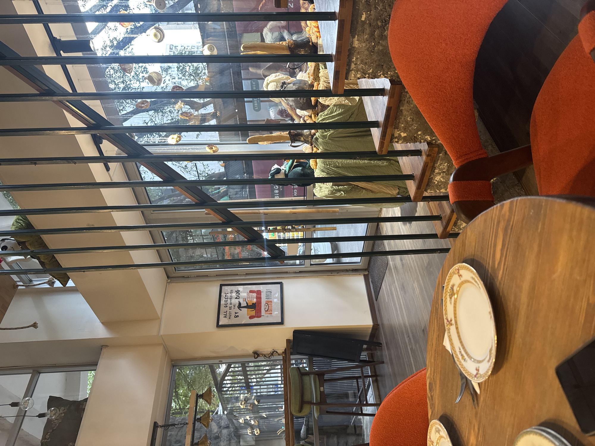 The image shows the interior of a cafe, likely in Tbilisi, Georgia, with an emphasis on rustic and industrial design elements. In the foreground, a round wooden table is set with decorative plates and silverware, suggesting a dining area. Two vibrant orange chairs with unique curved designs are positioned around the table, adding a pop of color to the scene.
To the left, a large window reveals a glimpse of the outdoors, with trees and a hint of urban architecture visible. Inside, a green bar stool is placed by a counter, accompanied by a black chalkboard. Further back, a staircase with wooden treads and metal railings ascends, leading to an upper level. The staircase is partially obscured by hanging decorative items, possibly teacups or small ornaments, suspended by chains.
The cafe appears to have an eclectic decor, with framed artwork on the wall, including a humorous poster featuring a dog. Various objects are displayed on shelves and surfaces, contributing to a cluttered yet charming ambiance. The overall impression is of a cozy and stylish establishment that embraces a unique aesthetic.