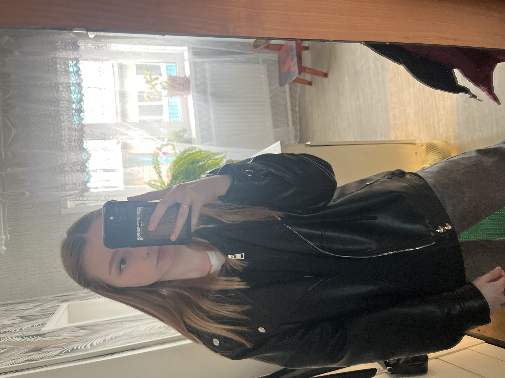 The image shows a selfie taken in a mirror in a room. The person taking the photo appears to be a fair-skinned young woman with long brown hair, wearing a black leather jacket over a white shirt. She's holding a smartphone with a black case.
The reflection in the mirror reveals details of the room. There's a window with sheer white curtains, revealing daylight and foliage. A child's chair is visible in the background. Part of a wooden frame is seen in the mirror's edge. The wallpaper visible near the mirror has a light-colored pattern.
Based on the location specified, the photo was likely taken indoors somewhere in Chaikovsky, Russia.