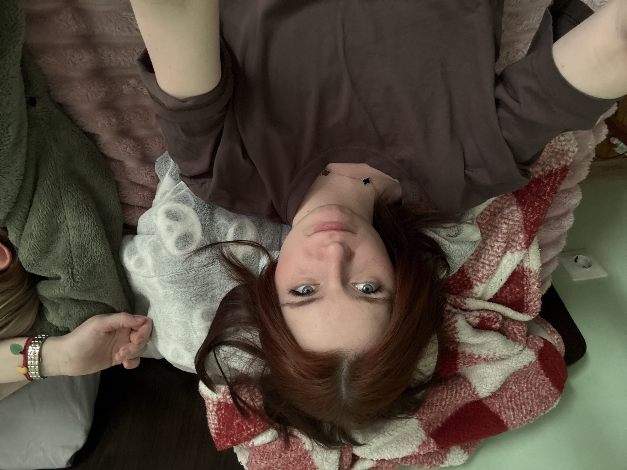 A woman is lying on a bed, looking up at the camera. She has reddish-brown hair, fair skin, and blue eyes. She is wearing a brown long-sleeved shirt and a silver necklace with clover-shaped pendants. She is surrounded by blankets and pillows of various colors and textures, including a green blanket, a pink blanket, a gray blanket with a pattern, and a red and white checkered blanket. A hand wearing bracelets is visible in the lower left corner. A wall outlet is visible in the upper right corner. The location is Unknown, Unknown.